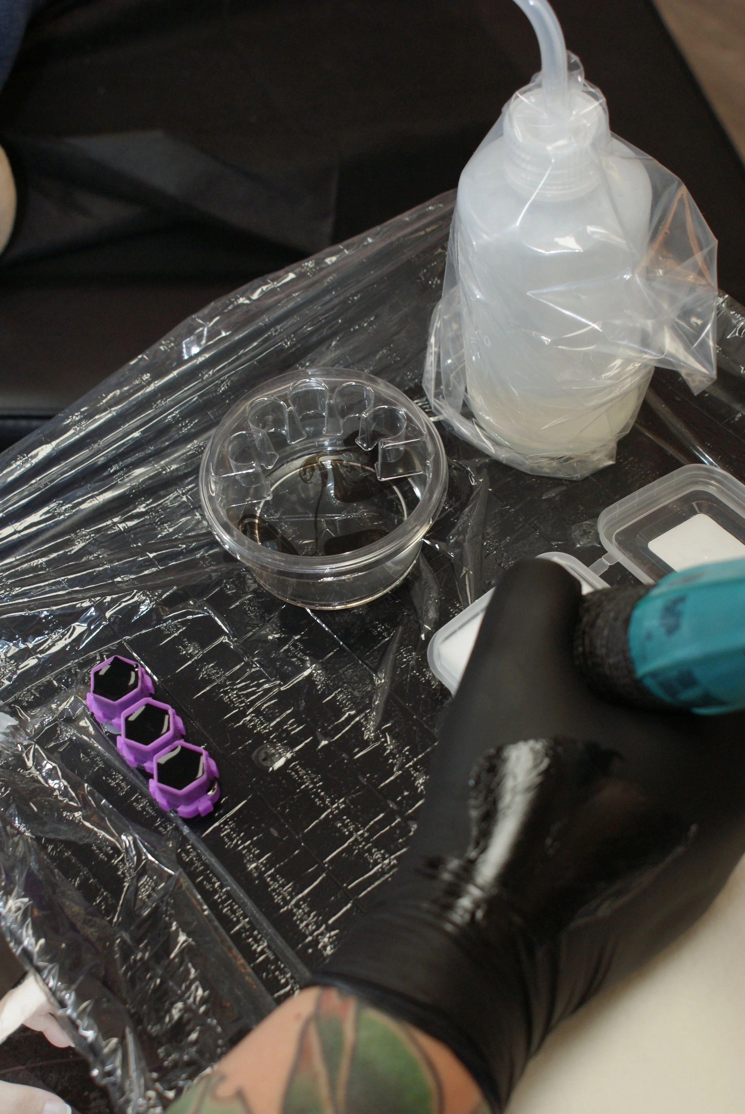 Close-up of tattooed arm, tattoo features a green and black design, placed above a person working with plastic tools and containers, on a black surface covered with aluminum foil.