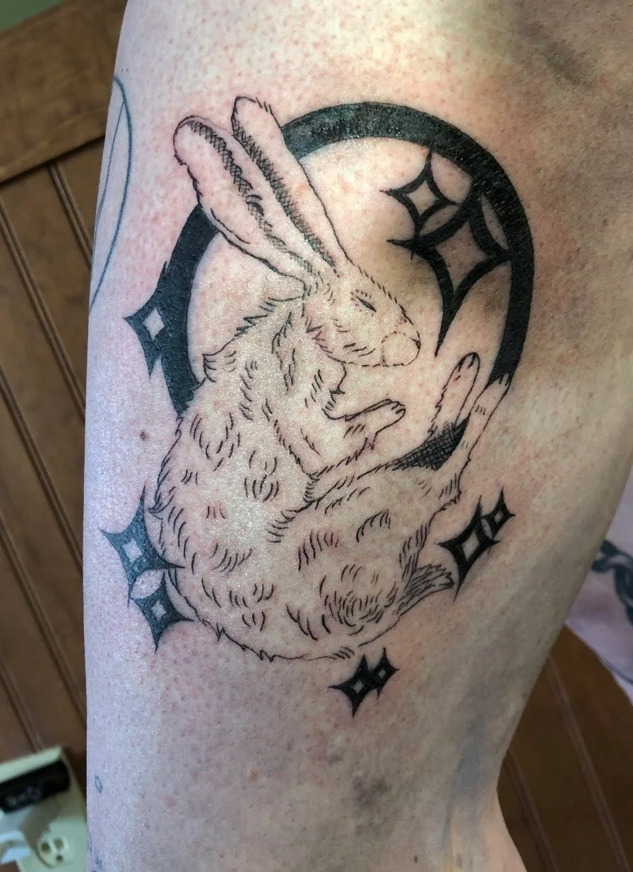 Black line tattoo of a rabbit with detailed fur, crouched inside a circle surrounded by star-shaped sparkles on a person's skin.