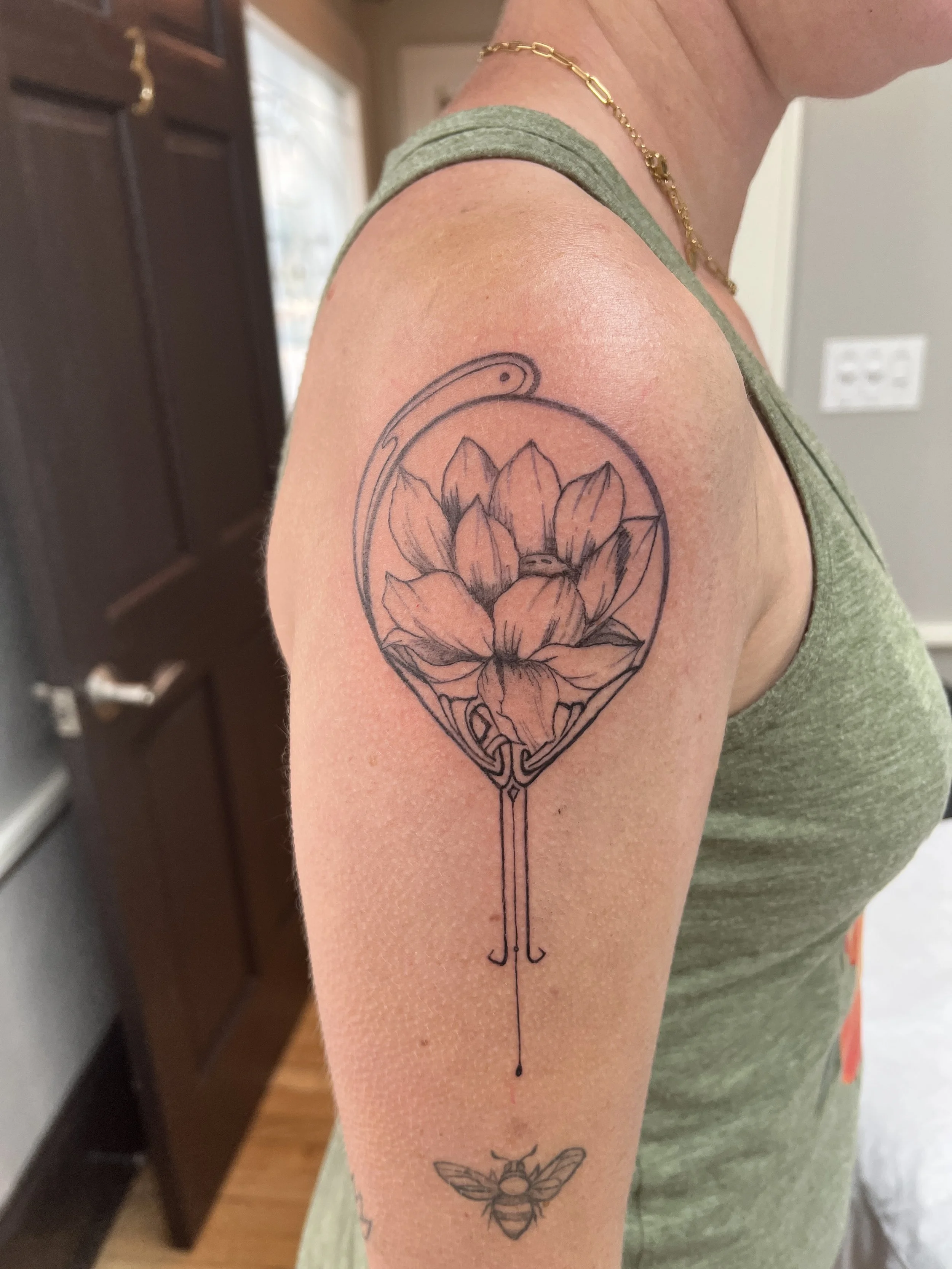 A person's upper arm with a floral tattoo inside a circle, a bee tattoo below, and a hanging umbrella and leaf tattoo lower on the arm. The person is wearing a green tank top, a gold chain, and a bracelet.