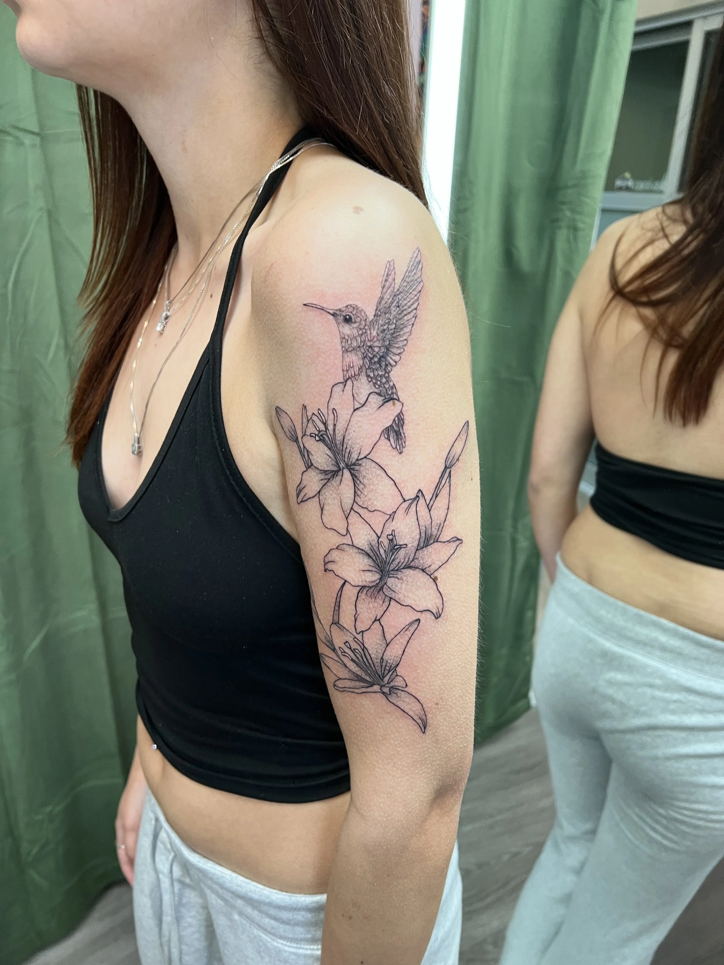 Close-up of a woman's upper arm and shoulder with a tattoo of a hummingbird and flowers. The woman is wearing a black tank top and layered necklaces.