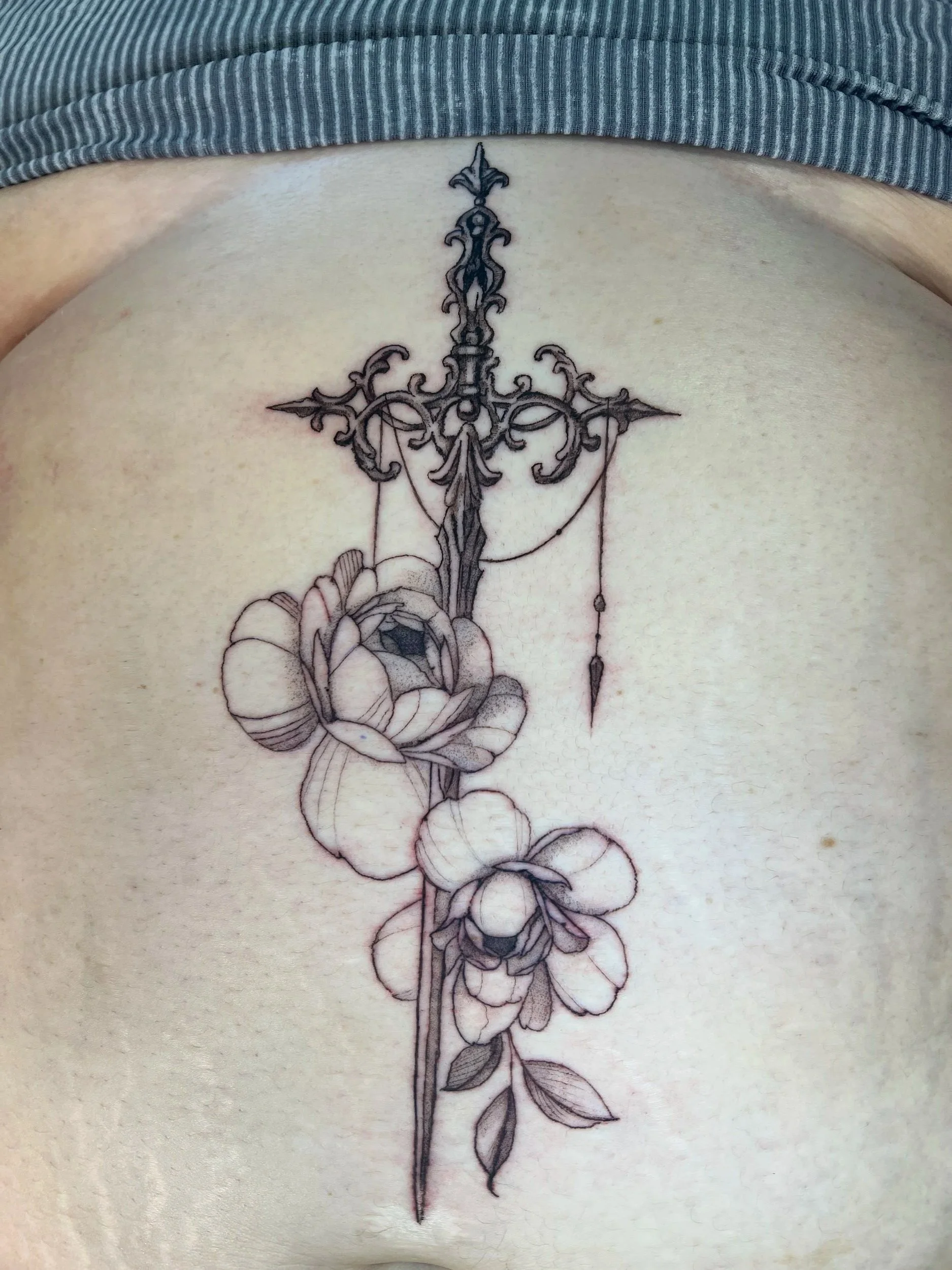 A black and gray fine line art tattoo of a sword with a decorative hilt and a rose along the length of the blade.