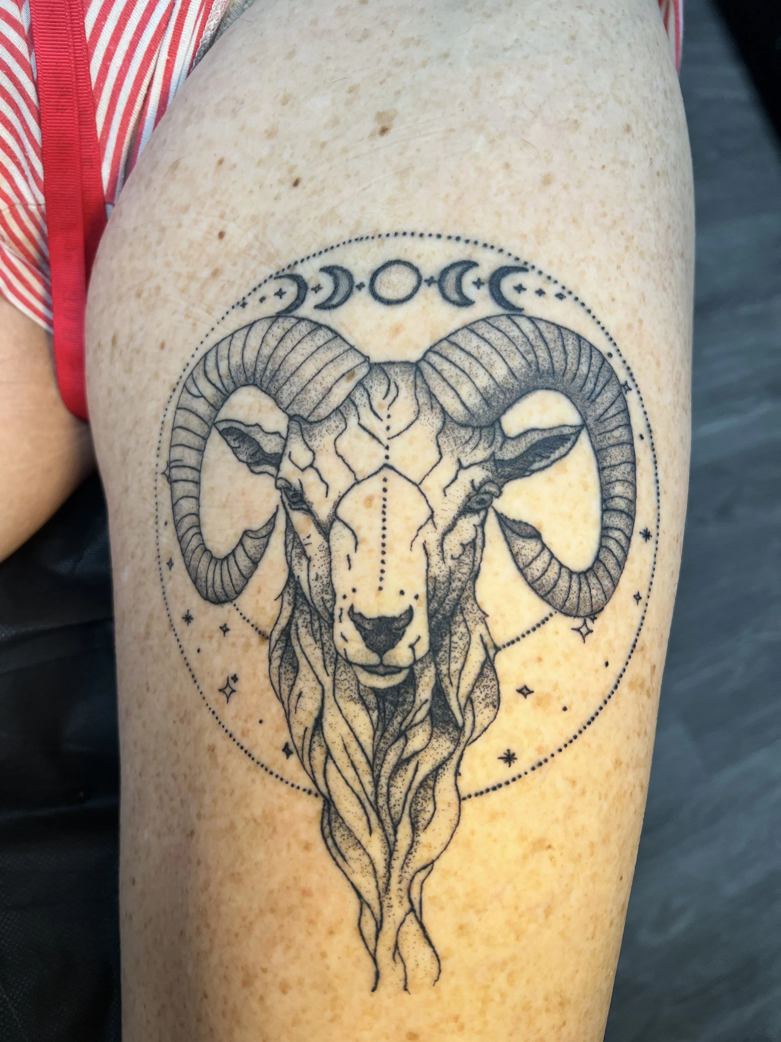 Tattoo of a ram's head with large curved horns, surrounded by celestial symbols including moons and stars, on a person's upper arm.