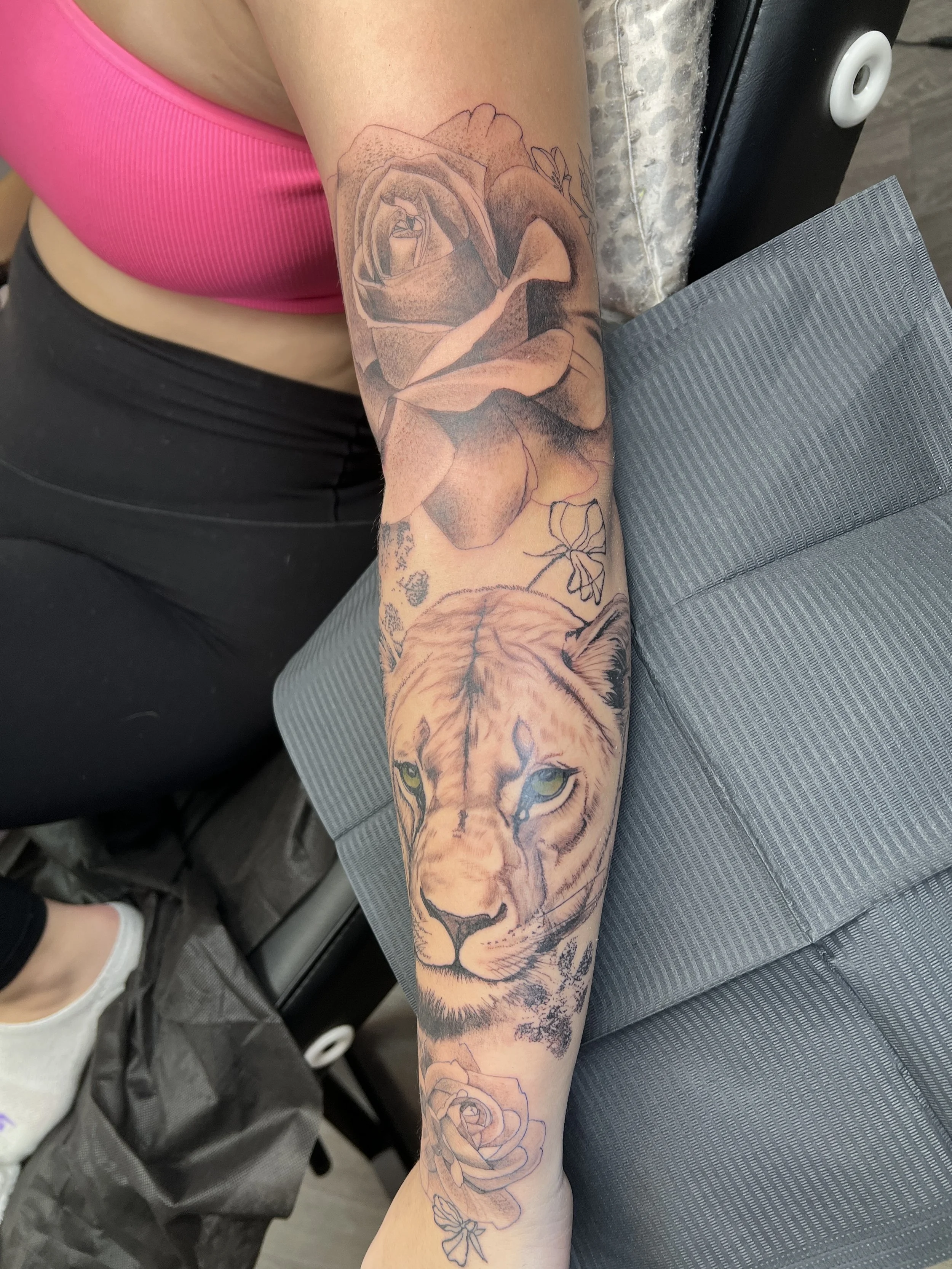Tattooed arm with a detailed realistic tiger face and a large flower, possibly a rose, on the forearm, sitting on a gray chair with a tattoo pad nearby.
