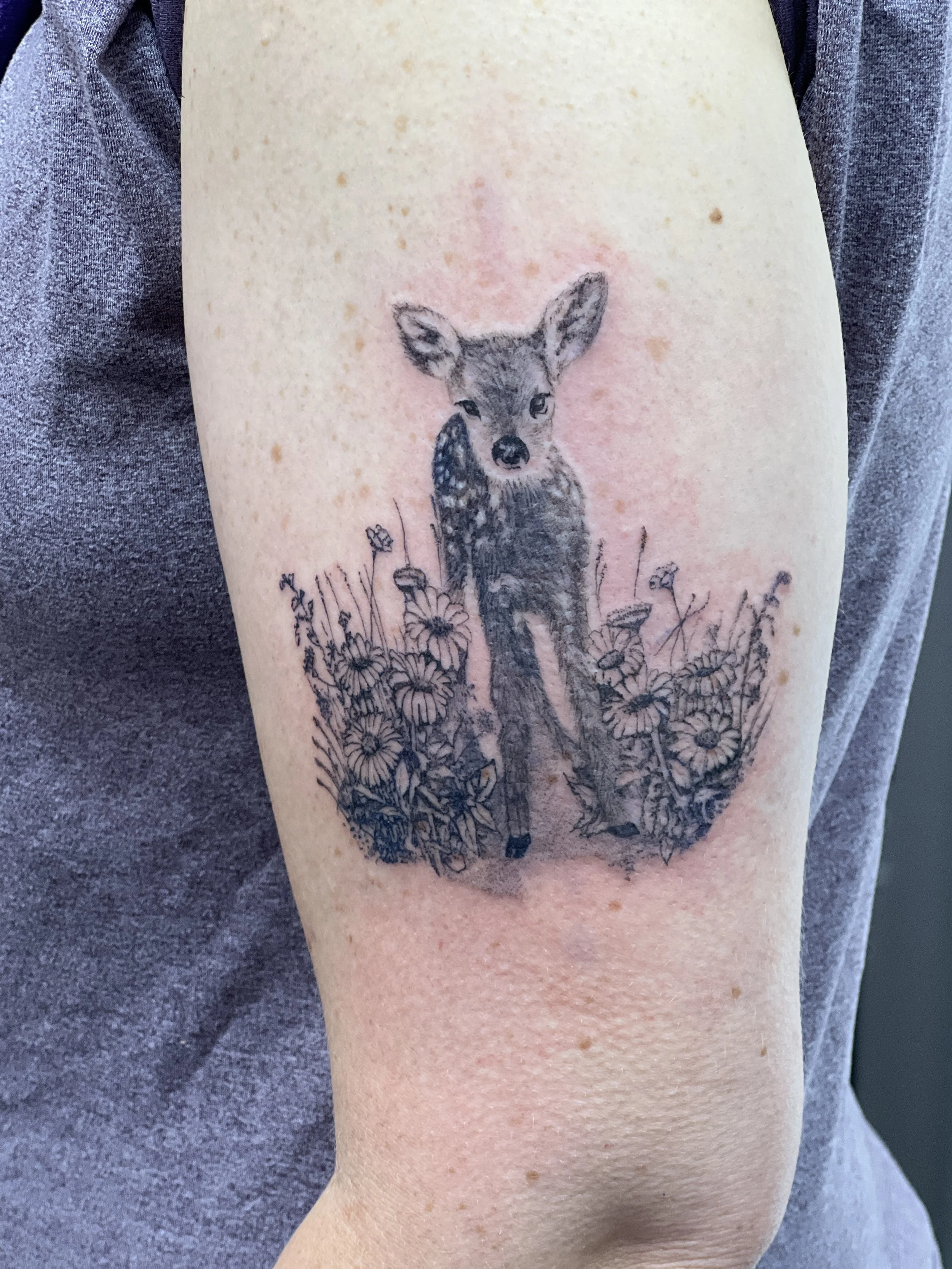 Tattoo of a deer with floral and grass details on a person's upper arm.