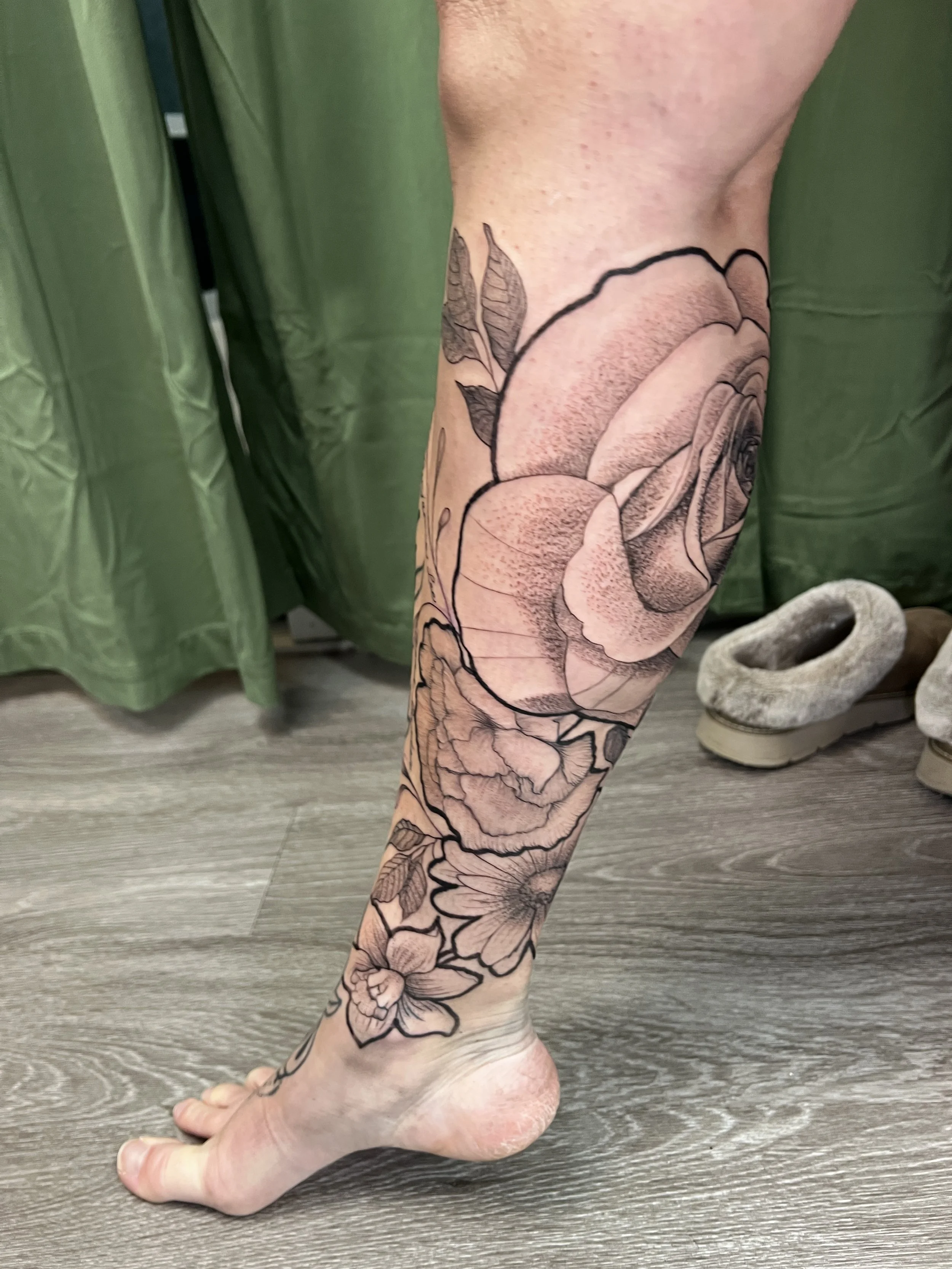 A person's leg with a large floral tattoo extending from the ankle up to the thigh, features outlined flowers and leaves.