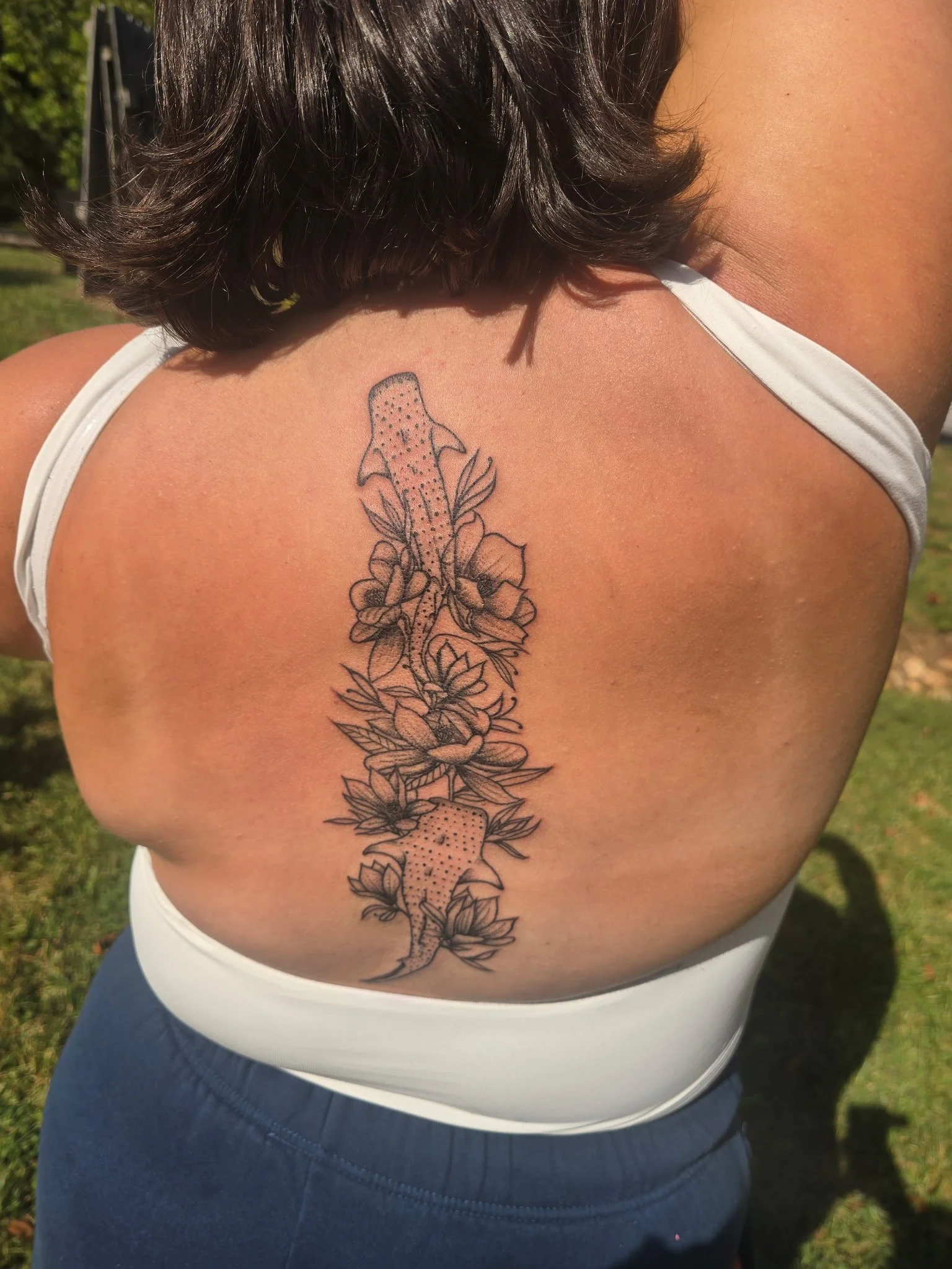 A person with dark hair wearing a white tank top and dark pants, showing a tattoo of two salamanders surrounded by flowers on their upper back near the shoulder.