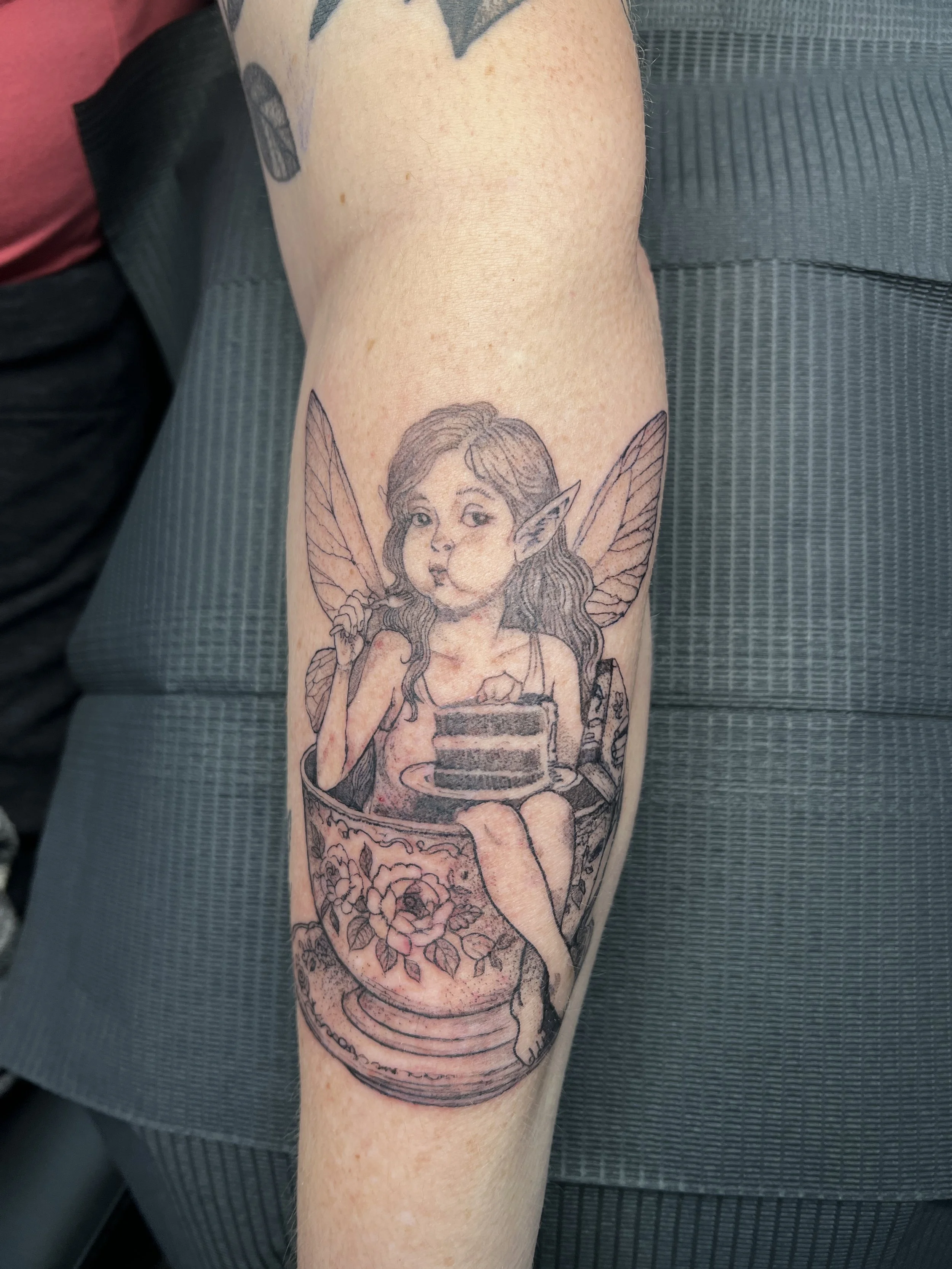 Tattoo of a fairy with butterfly wings holding a cup, surrounded by flowers on a person's arm.