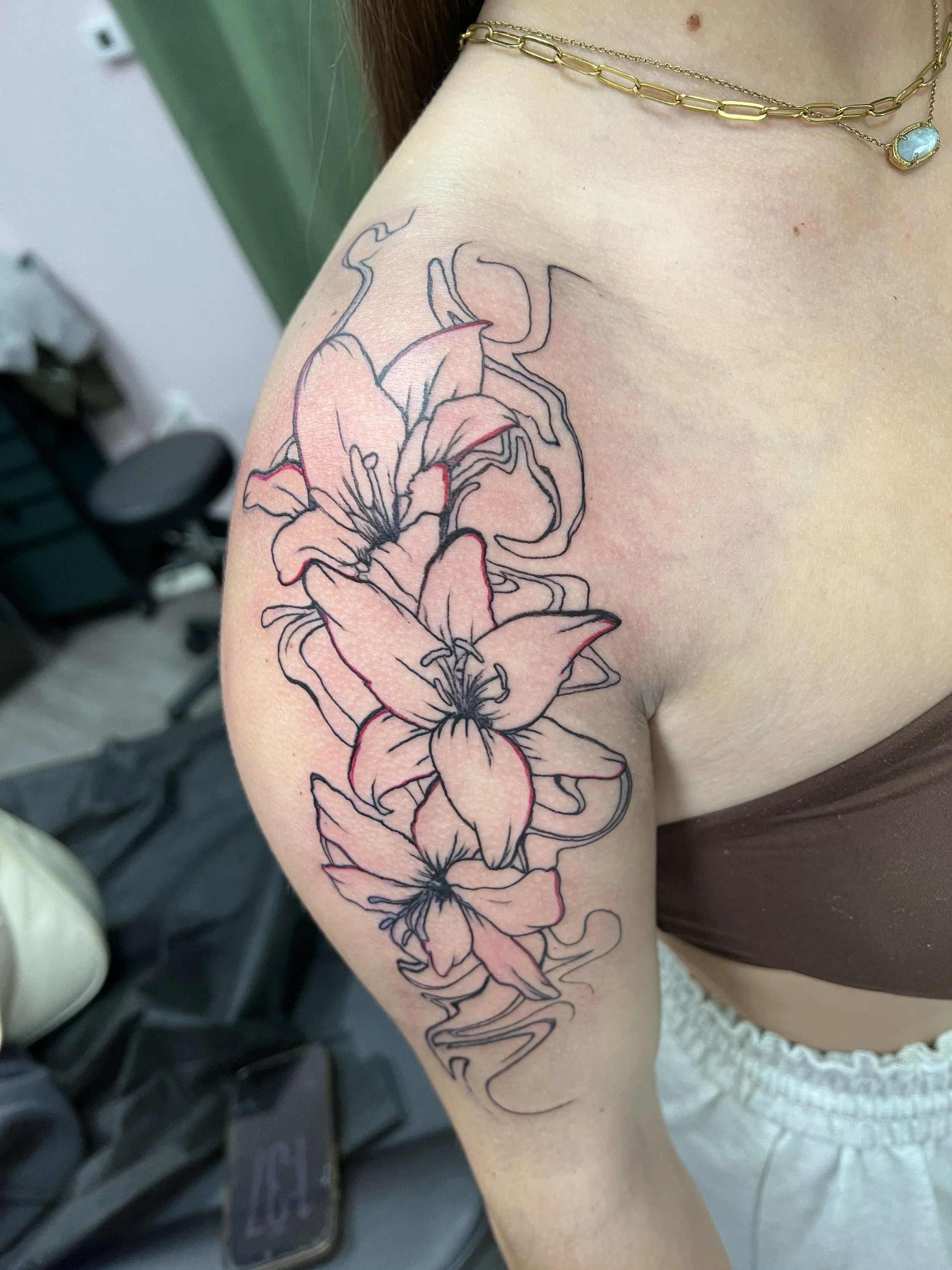 A tattoo of a large lily flower with detailed petals on a person's upper arm or shoulder. The person is wearing layered gold necklaces and brown clothing.