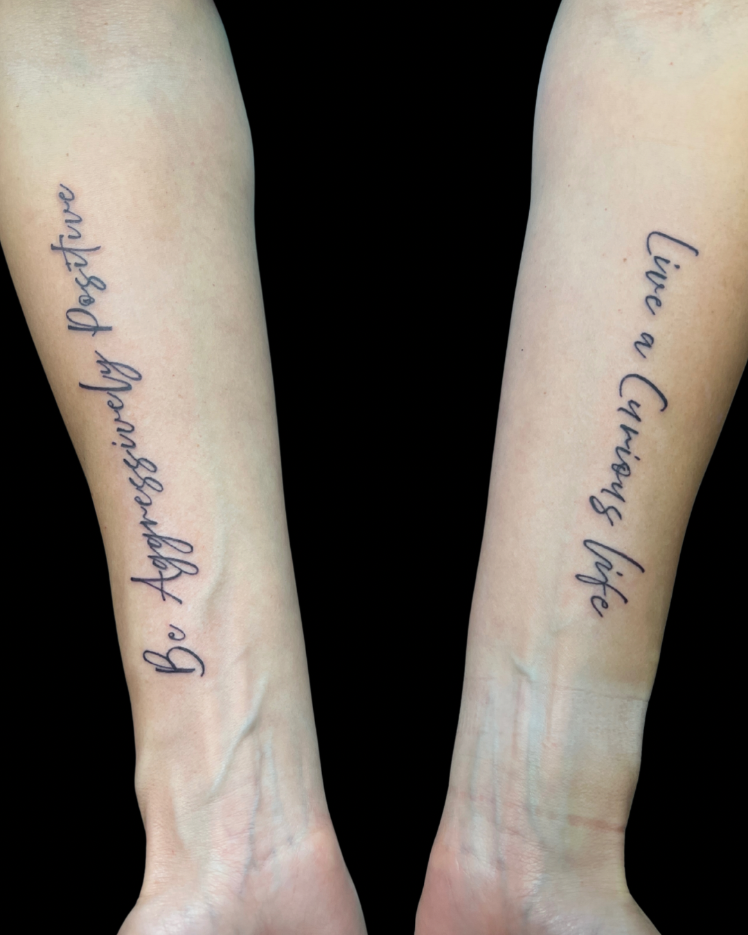 Two forearms with matching tattoos in cursive script: "Be Absolutely Postive" on the left arm and "Live a Champion" on the right arm, set against a black background.