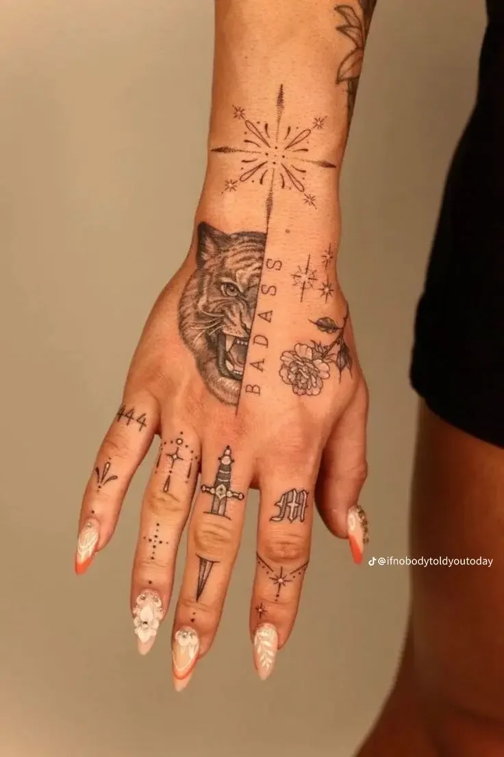 A person's hand with multiple tattoos, including a tiger face on the palm, the word 'BADASS' vertically on the middle finger, fireworks on the wrist, a rose and a leaf near the middle finger, and various smaller tattoos on the fingers.