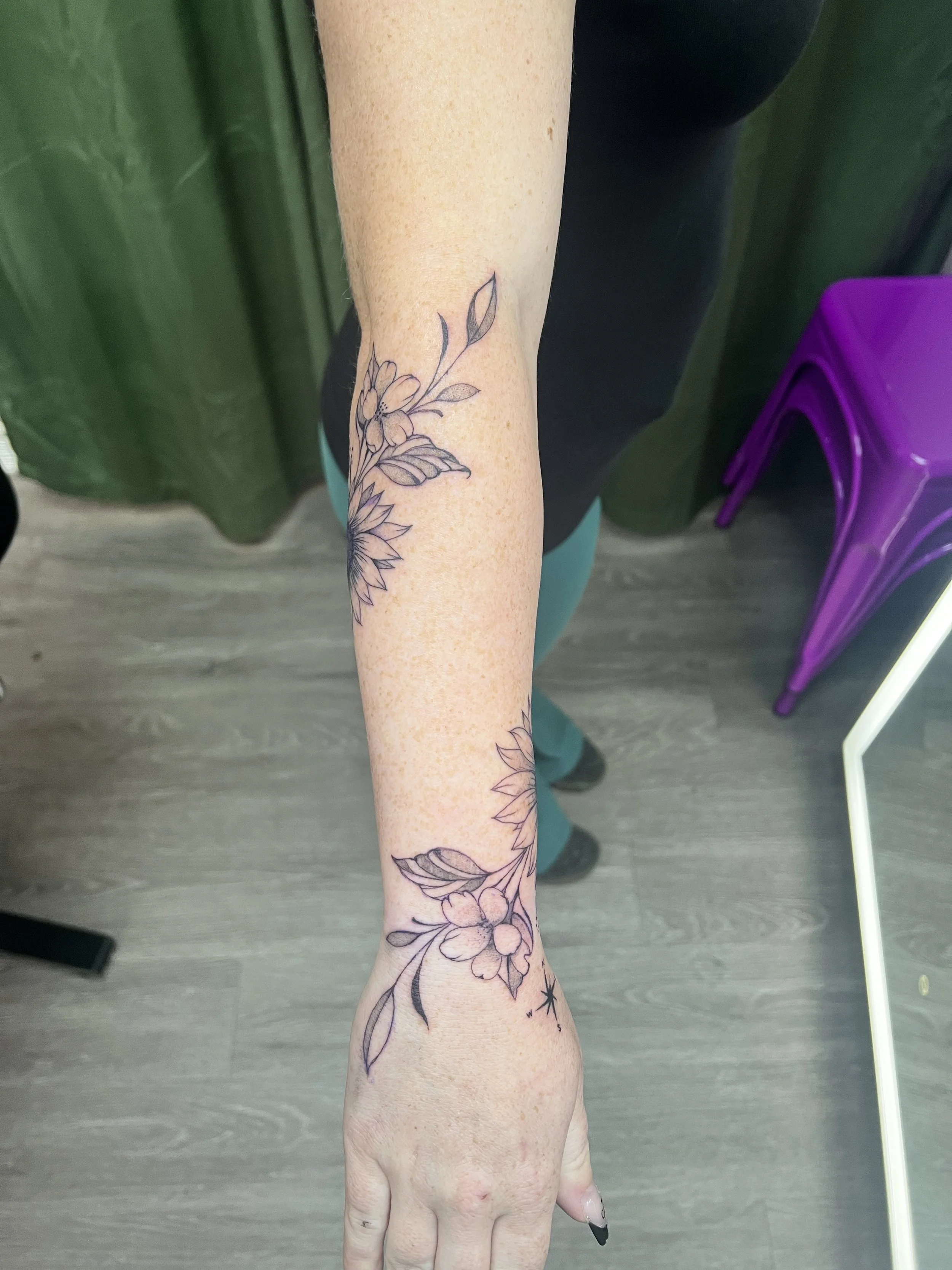 A tattoo of flowers and leaves on a person’s forearm and hand, with a star near the wrist. The person is wearing black pants and a black top, with a green background and purple chair visible.