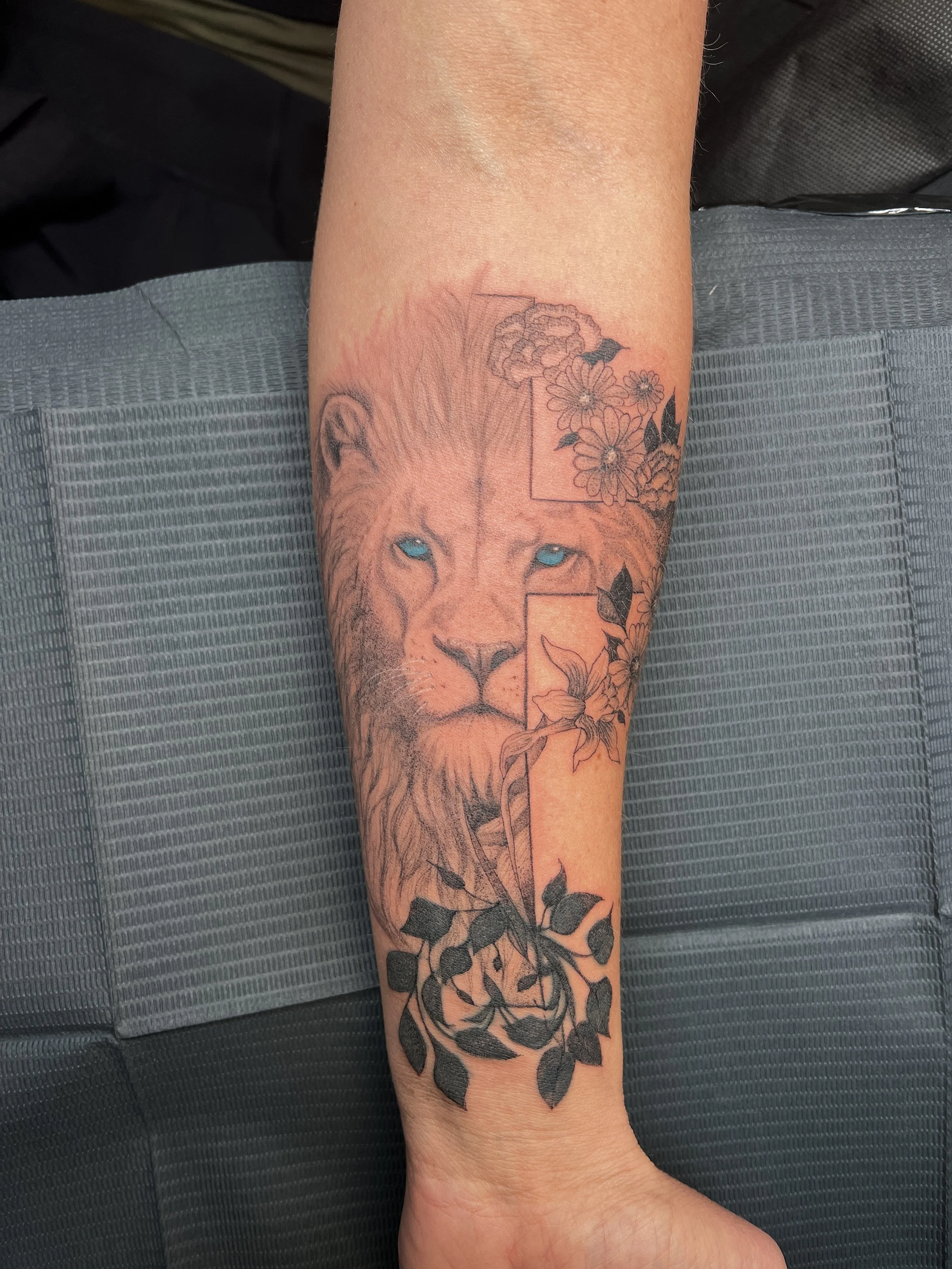 A tattoo on a person's forearm depicting a lion's face with light blue eyes, surrounded by flowers and leaves, with some flowers and leaves incorporated into a geometric square outline.