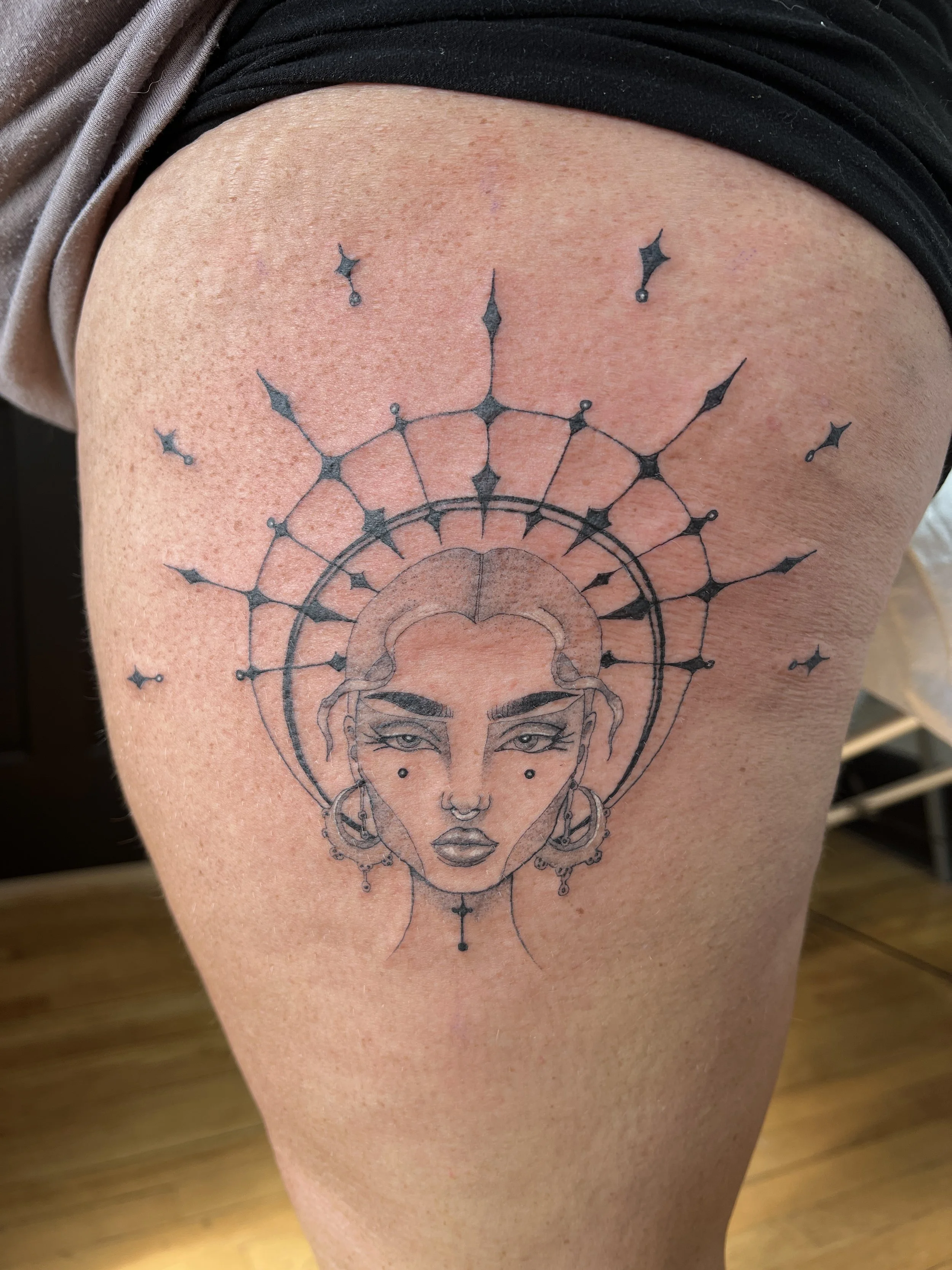 A tattoo of a woman's face with intricate geometric and celestial designs around her head on a person's upper arm.