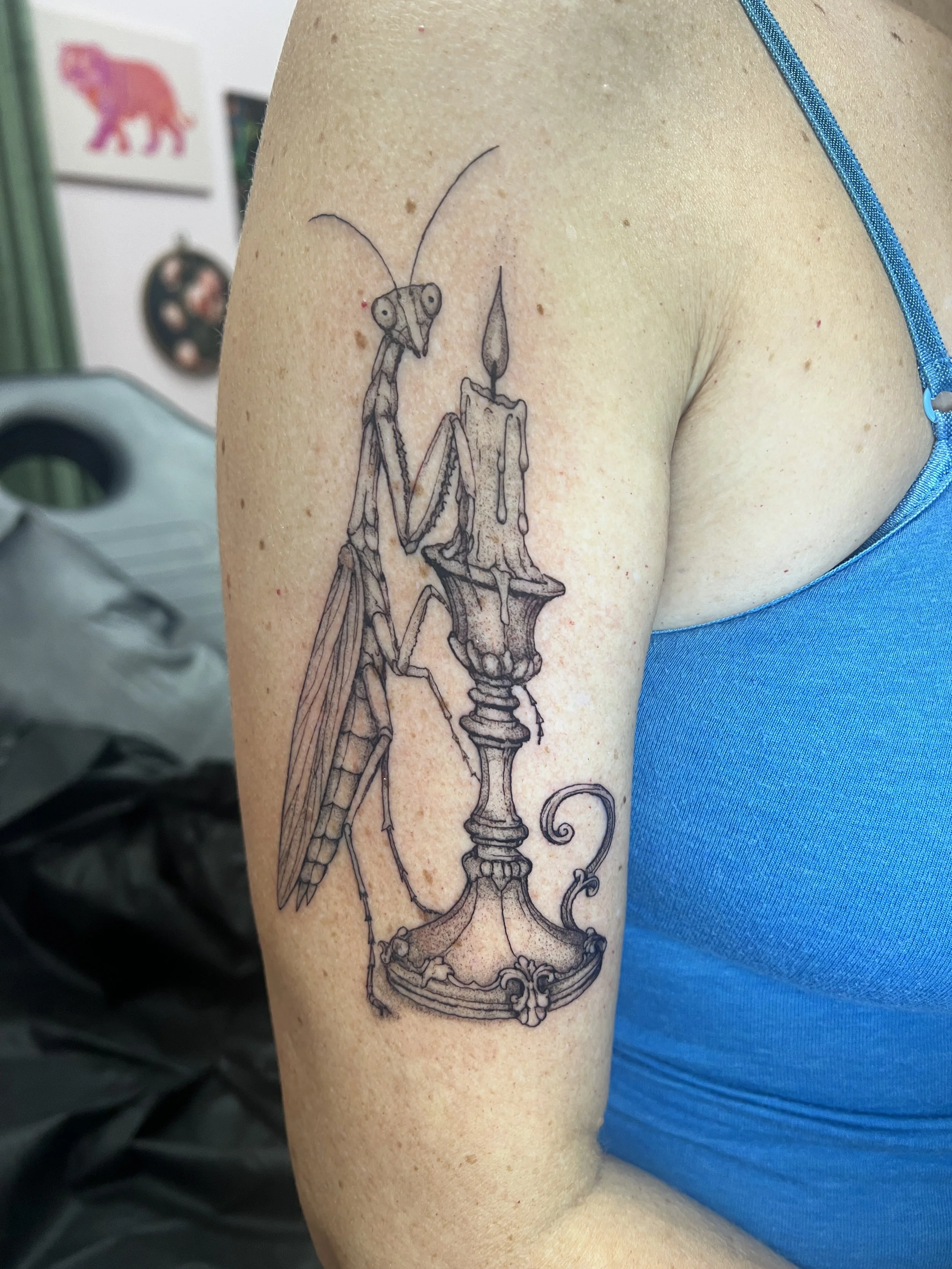 A tattoo of a praying mantis sitting on an ornate candle holder, inked on a person's upper arm.