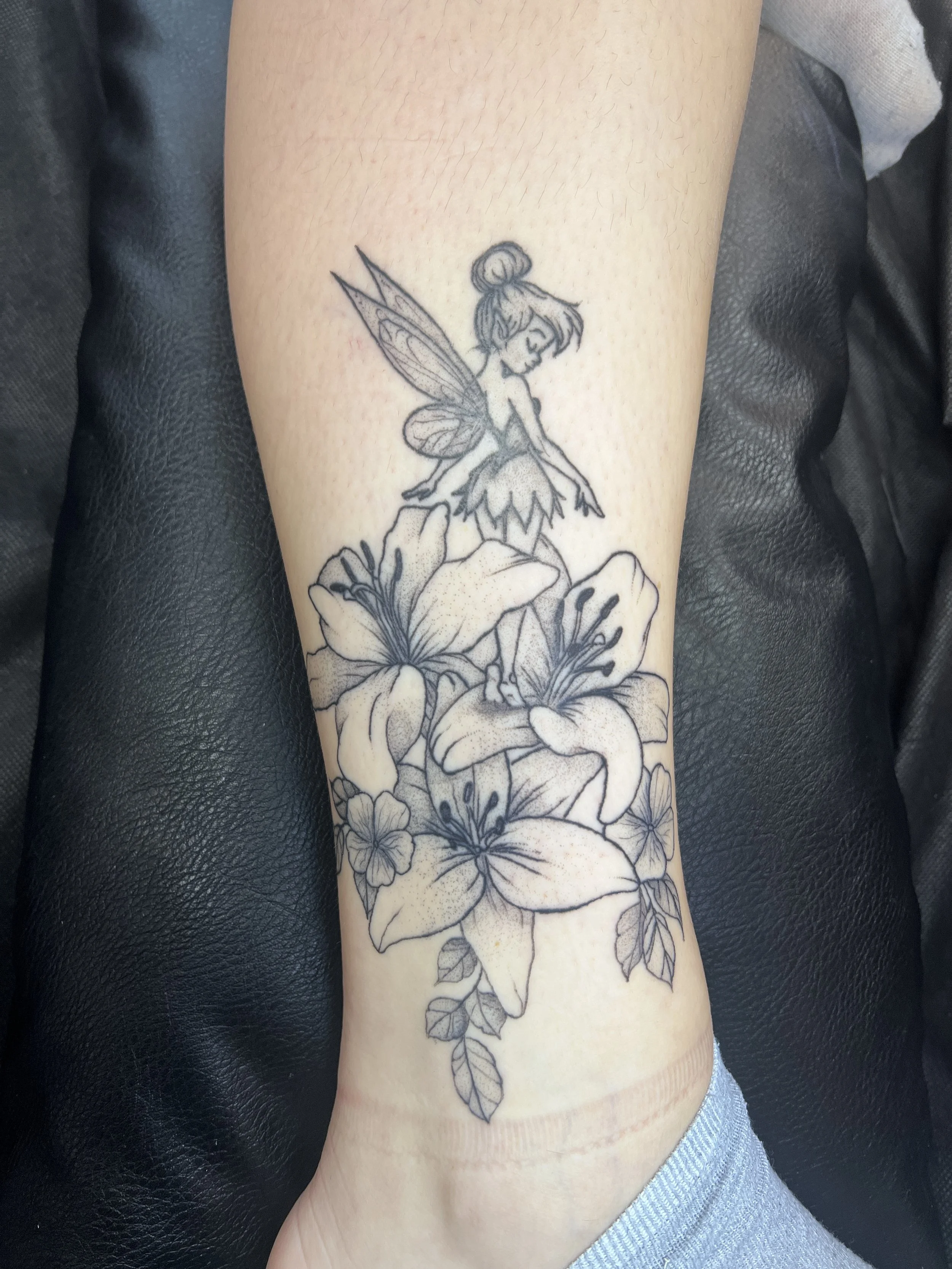 Black fine line tattoo of a fairy sitting on a flower, with large butterfly wings, on a person's forearm.