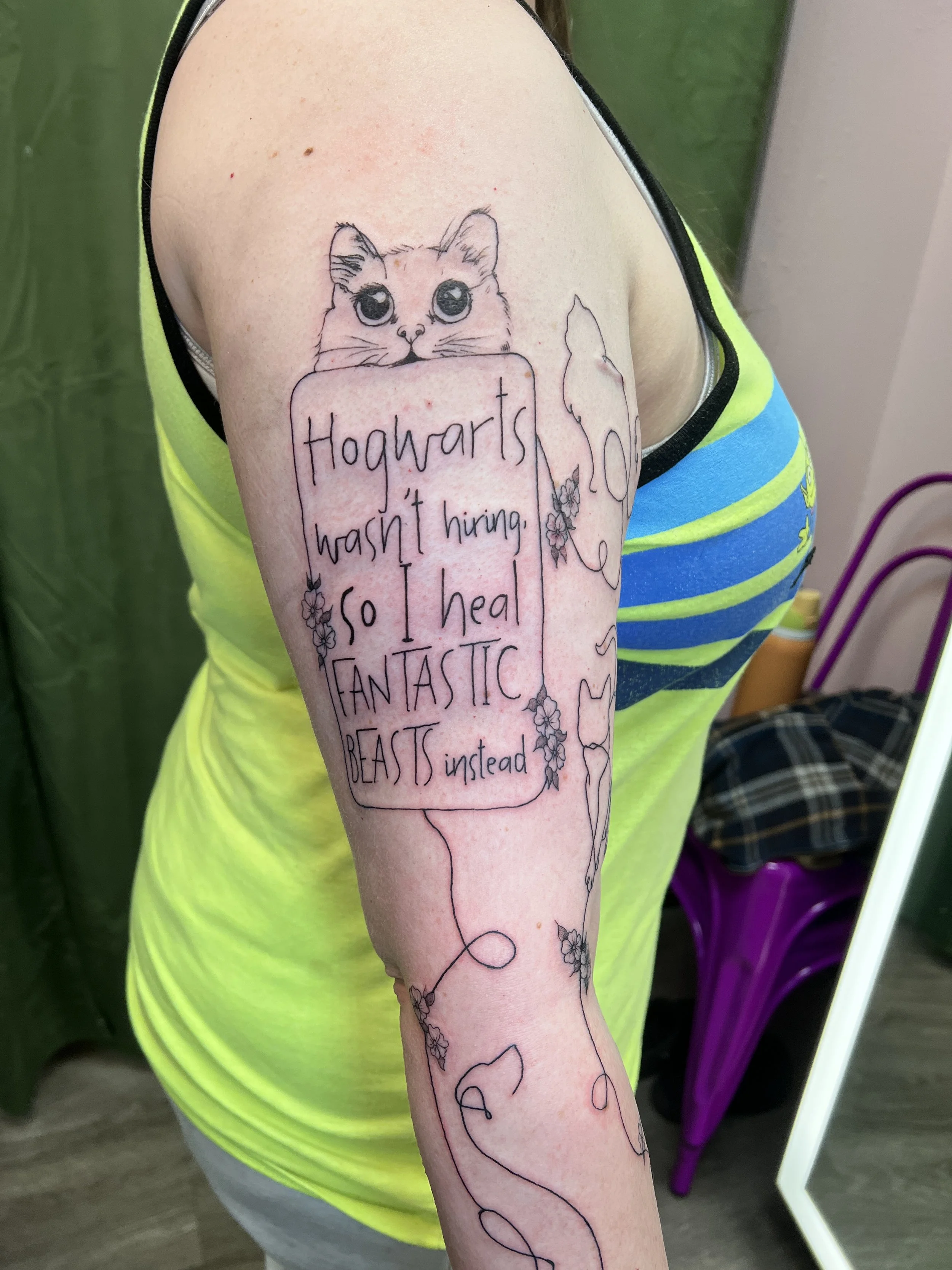 A person's arm with a fine tattoo of a cat peeking over a sign that reads 'Hogwarts hasn't hiring. So I call it FANTASTIC BEASTS instead.' The tattoo is outlined in black with minimal coloring, and there are small decorative flowers around the text.