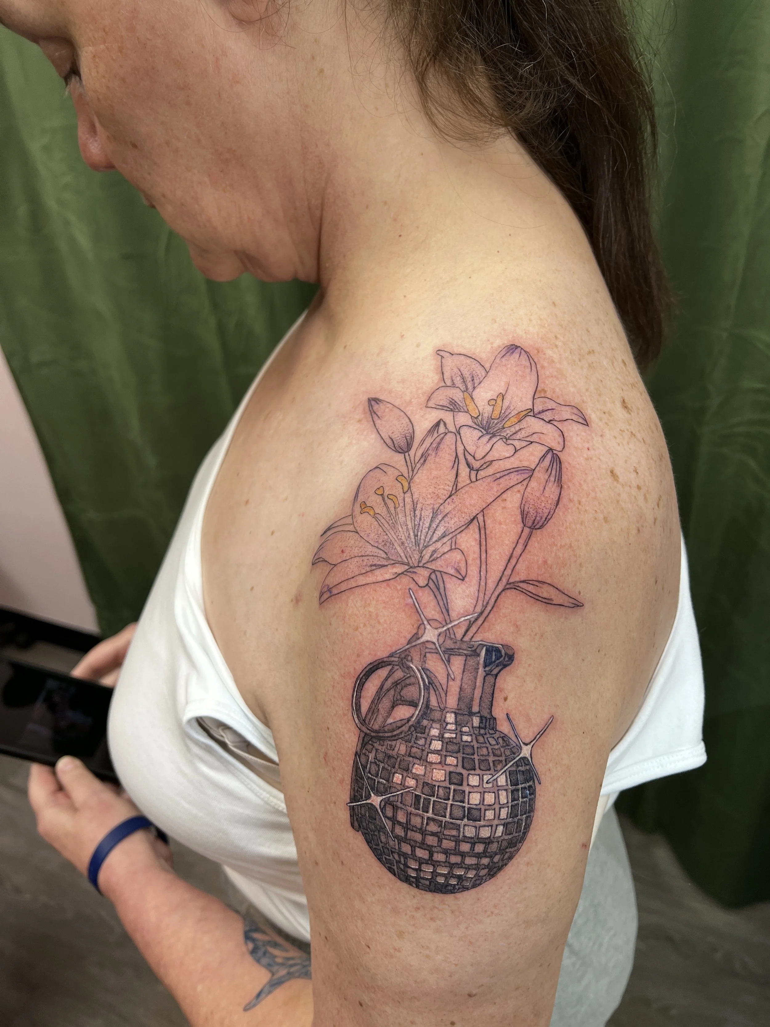 A woman with a tattoo of a flower and a skull on her upper arm, wearing a white sleeveless top, with the background of green leaves.