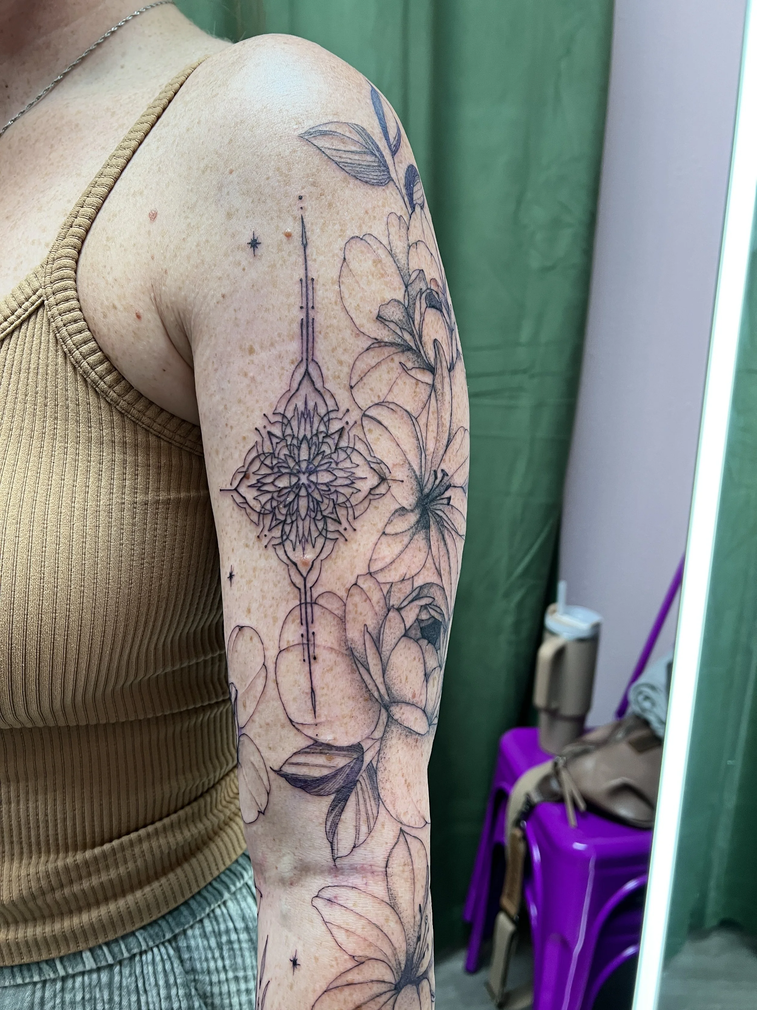 A person with a floral tattoo on their upper arm. The tattoo features flowers, leaves, and stars, displayed against a green and purple background.