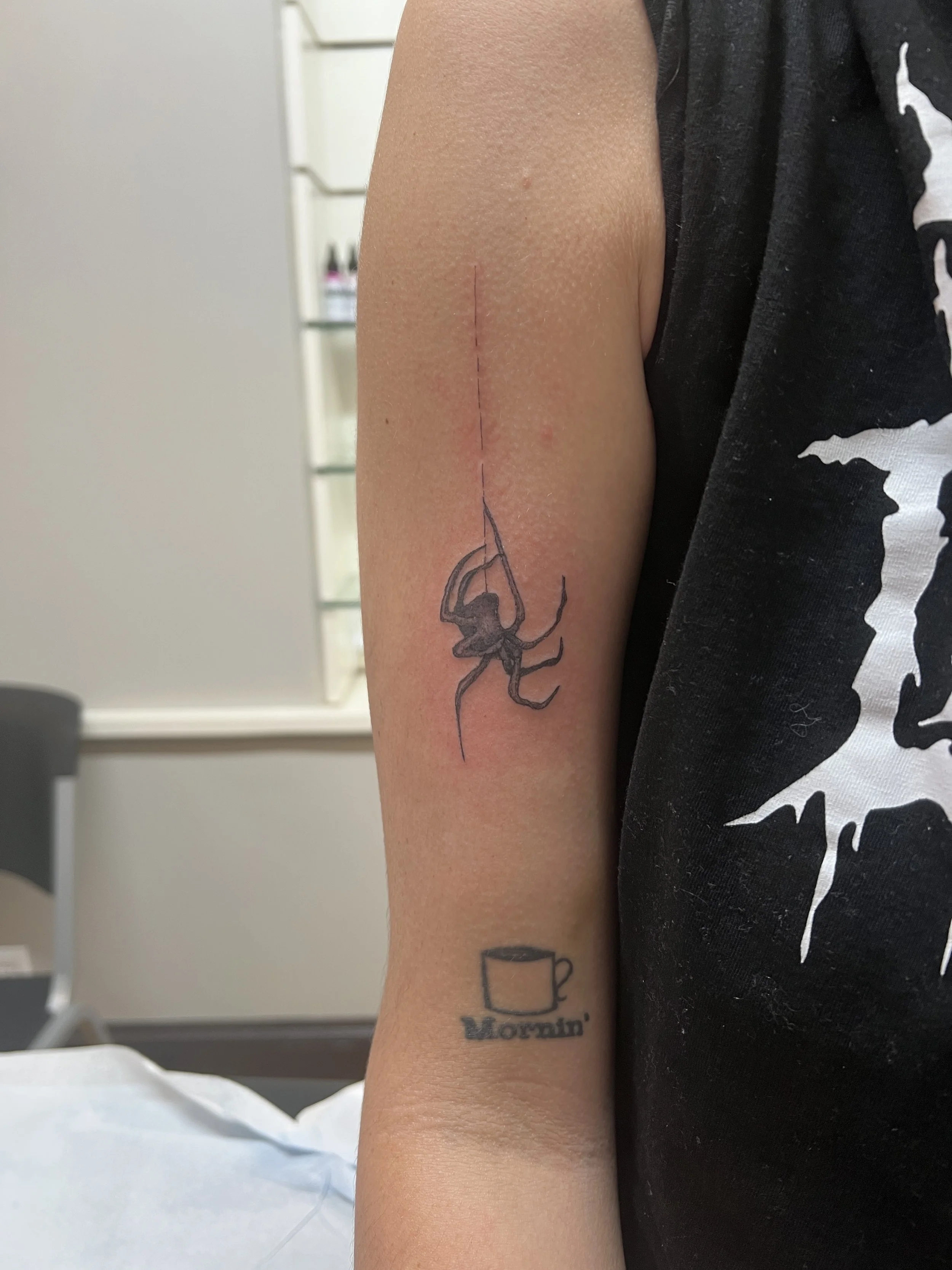 A person's arm with temporary tattoos, including a black outline of a spider and two cups, one with the word 'Yoursin' written underneath. The arm also has a fresh scratch.