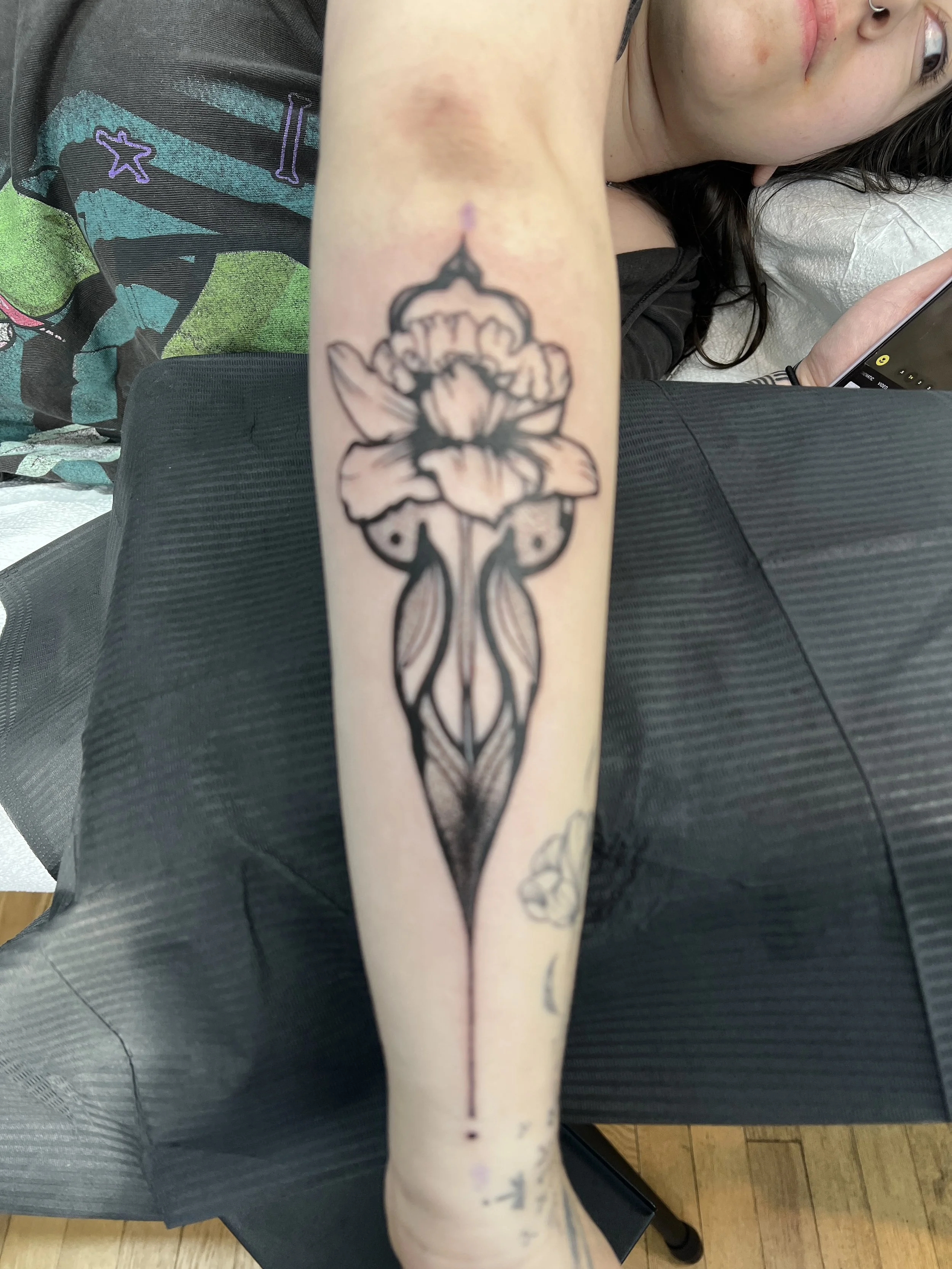 A person's inner forearm with a black outlined tattoo of a floral design and long, flowing stems. The tattoo is in black ink and located in the middle of the forearm.