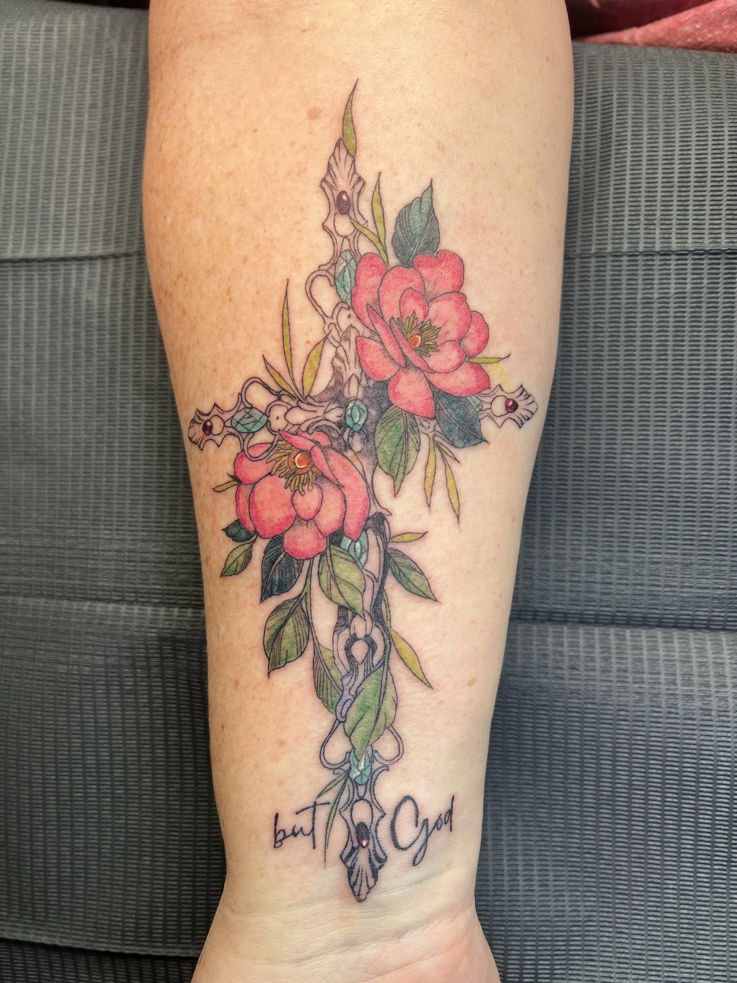 Colorful tattoo of flowers, leaves, and thorns on a person's arm with the words 'but God' written beneath.