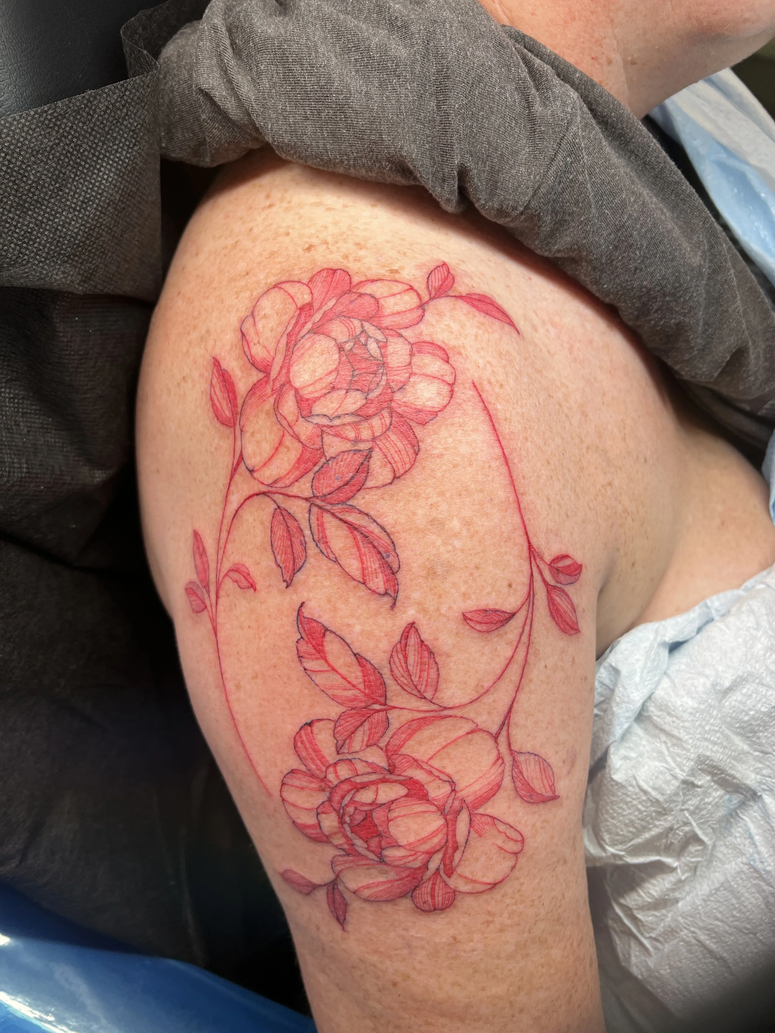 A person's upper thigh with a detailed outline of a rose tattoo in red ink, sitting on their lap or resting on a surface, wearing a dark gray shirt.