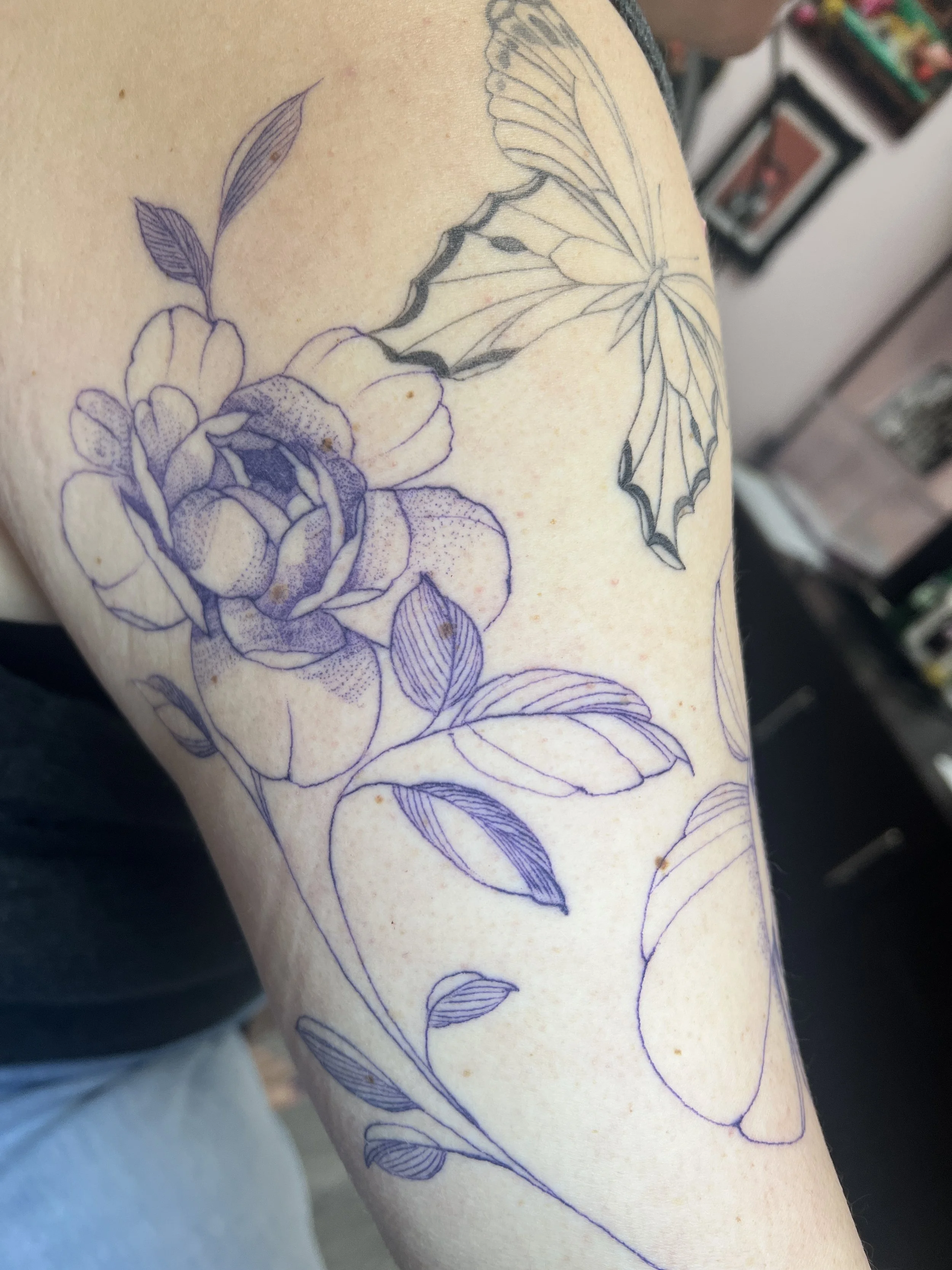A tattoo fine line of a flower and a butterfly on a person's arm, with the flower drawn in purple ink and the butterfly's wings outlined in black ink.