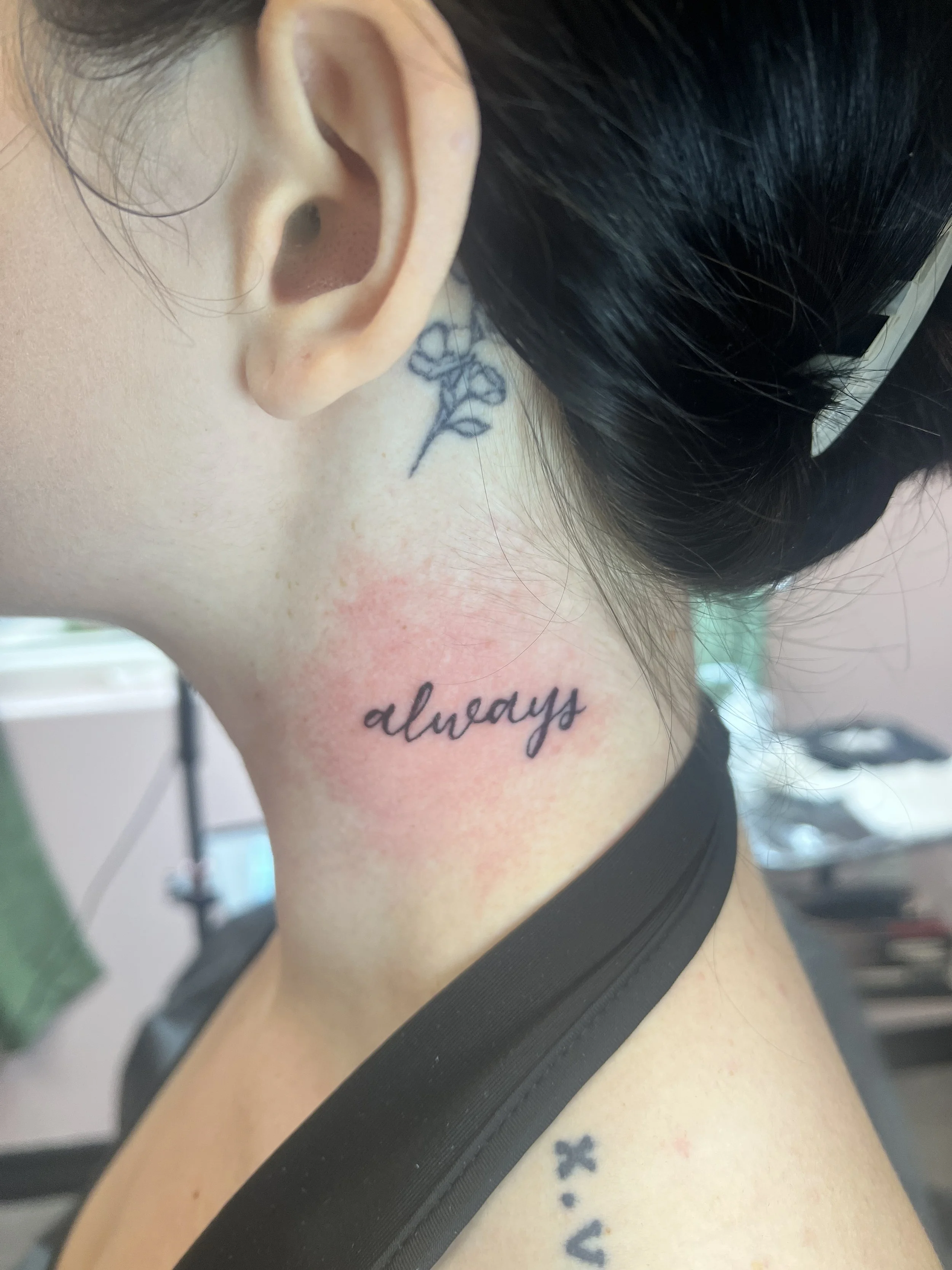 Close-up of a woman's neck with a tattoo that says 'always' and a small skull, as well as close-up of her ear and hair, with part of her face visible.