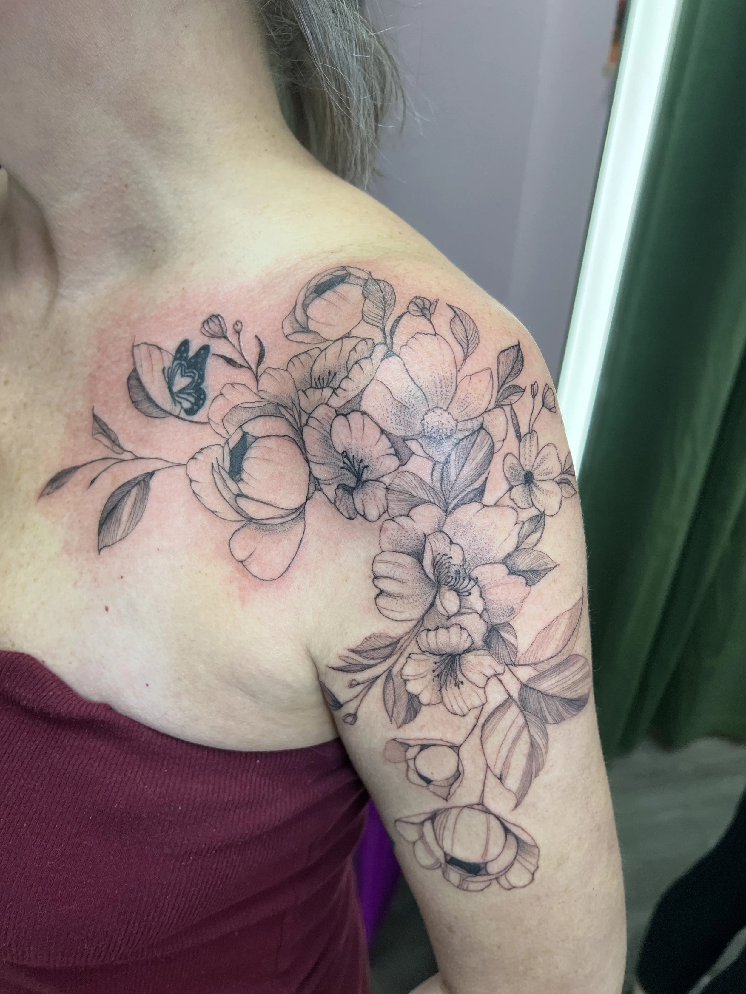 A close-up of a person's shoulder and upper arm with a floral tattoo featuring roses, leaves, and a butterfly, with some redness around the tattoo.