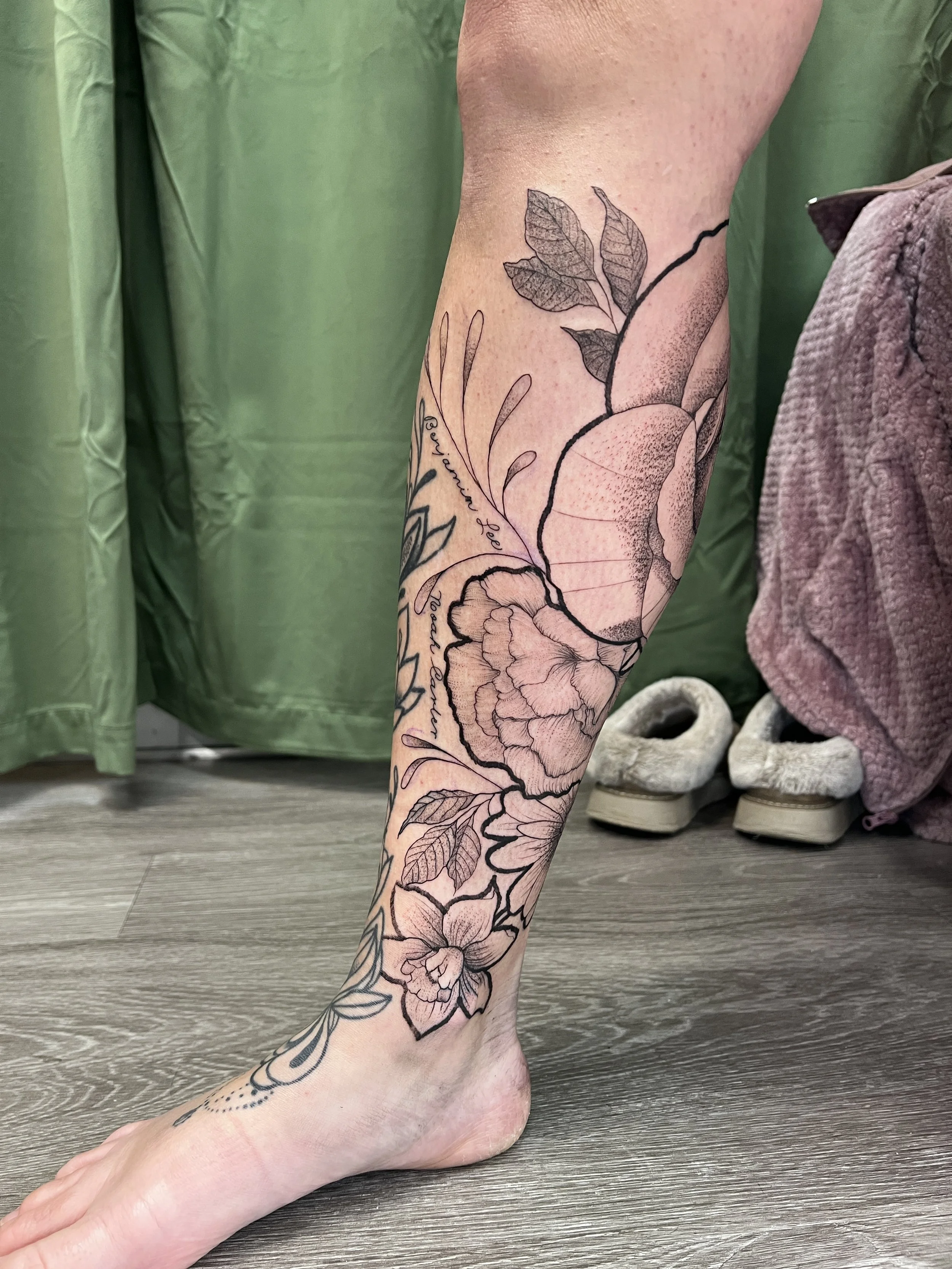 A person's lower leg and foot with a black ink floral tattoo featuring detailed leaves and flowers, set against a background of a wooden floor and green curtain.