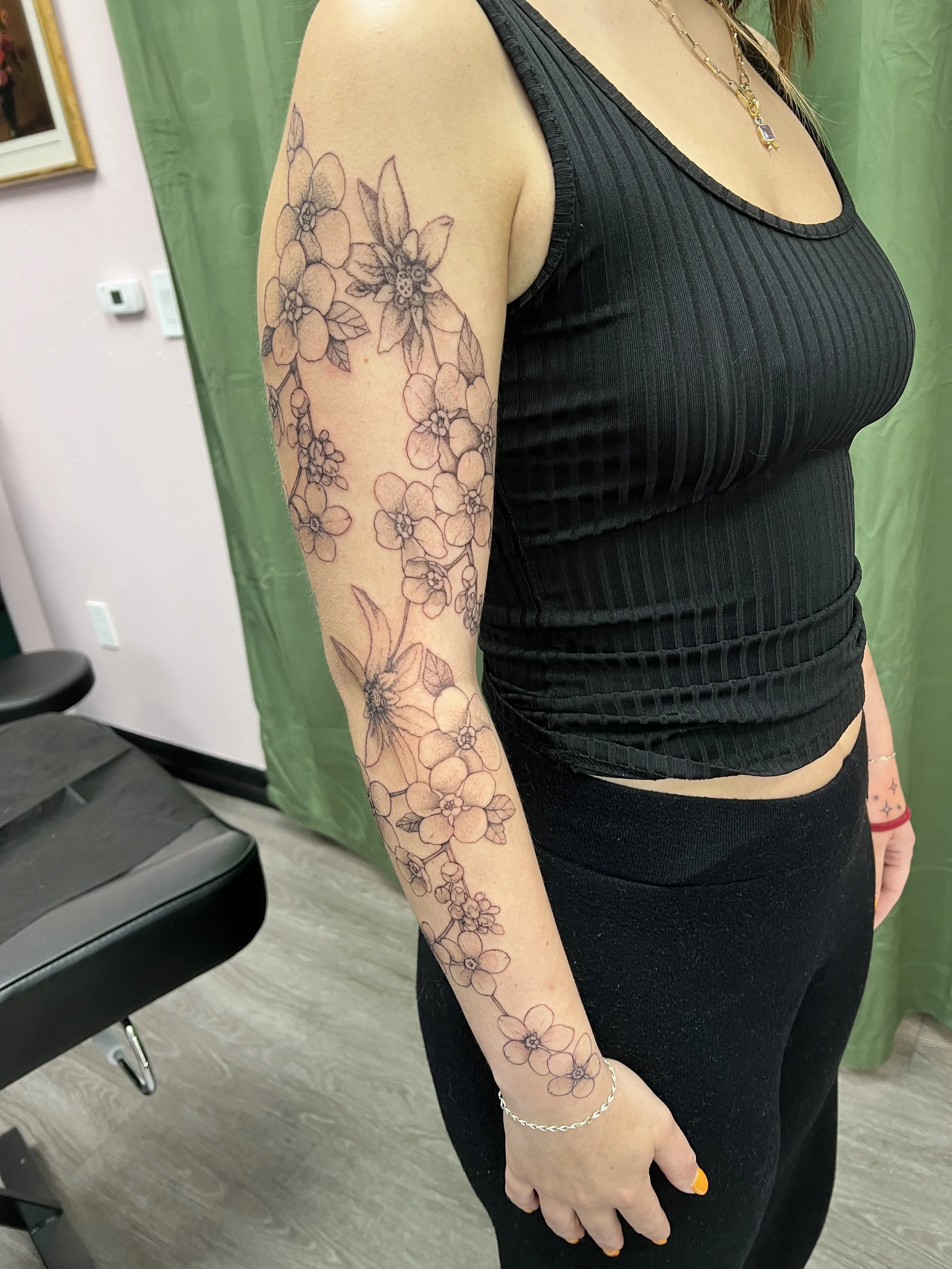 A woman with a floral fine line tattoo on her upper arm, wearing a black ribbed tank top, a gold necklace, and a silver bracelet, standing indoors with a green curtain and a framed picture in the background.