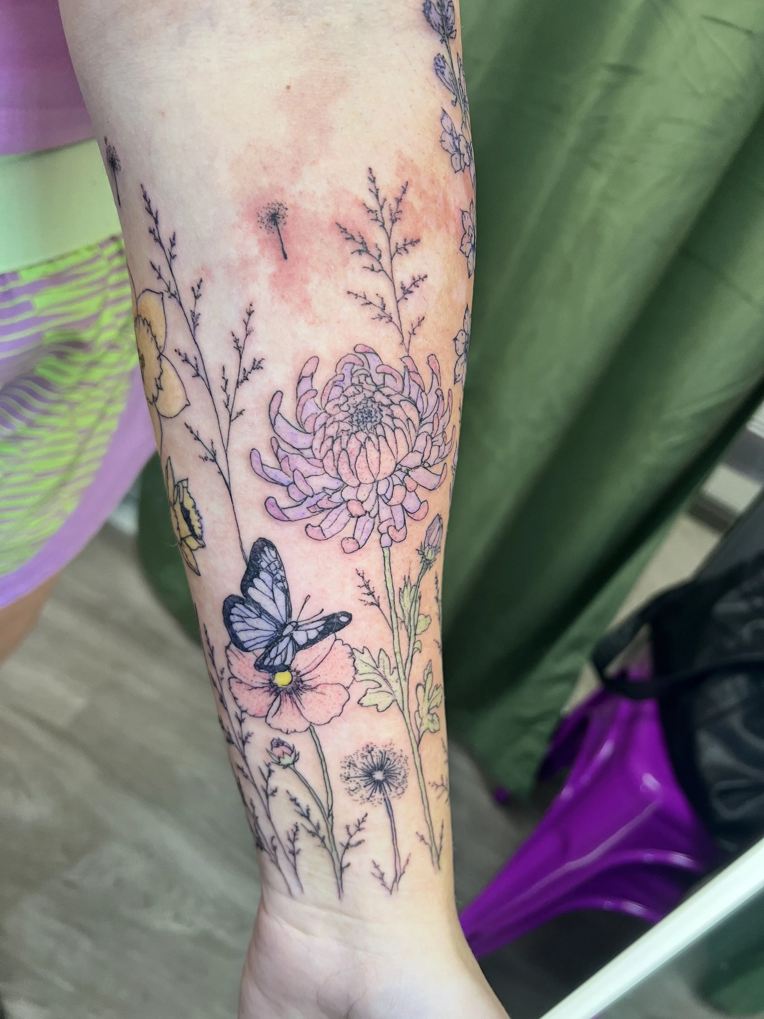 Close-up of a person's forearm with a floral tattoo featuring flowers, leaves, a butterfly, and dandelions.