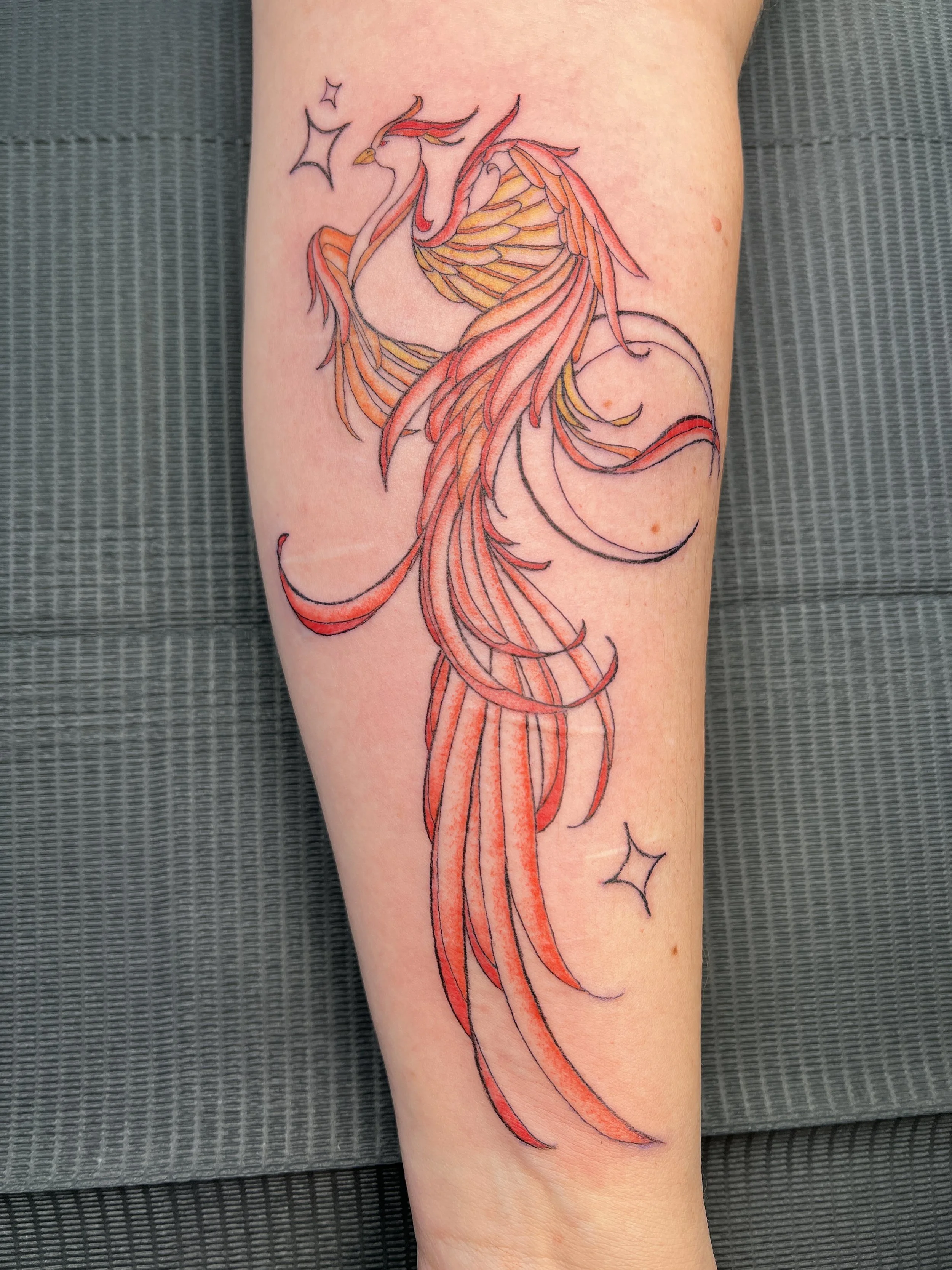 Colorful tattoo of a phoenix with detailed wings, flowing tail feathers, and stars around it on a person's body.