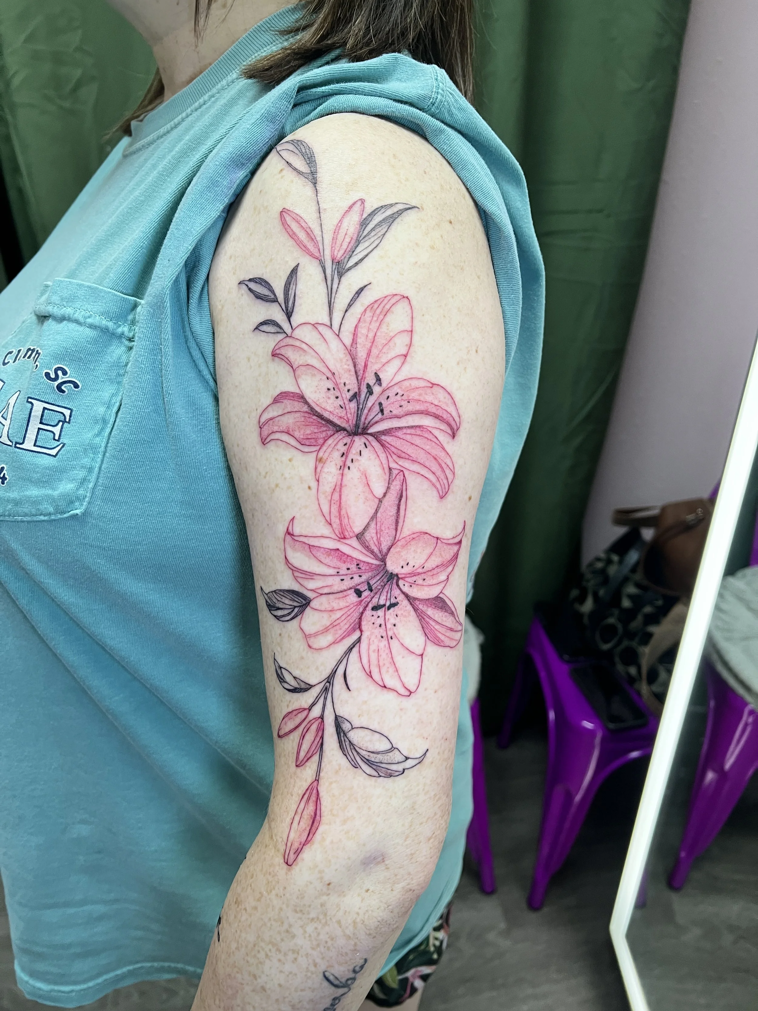 A person's upper arm with a floral tattoo featuring pink and black inked lilies and leaves, with some filled in and others outlined.