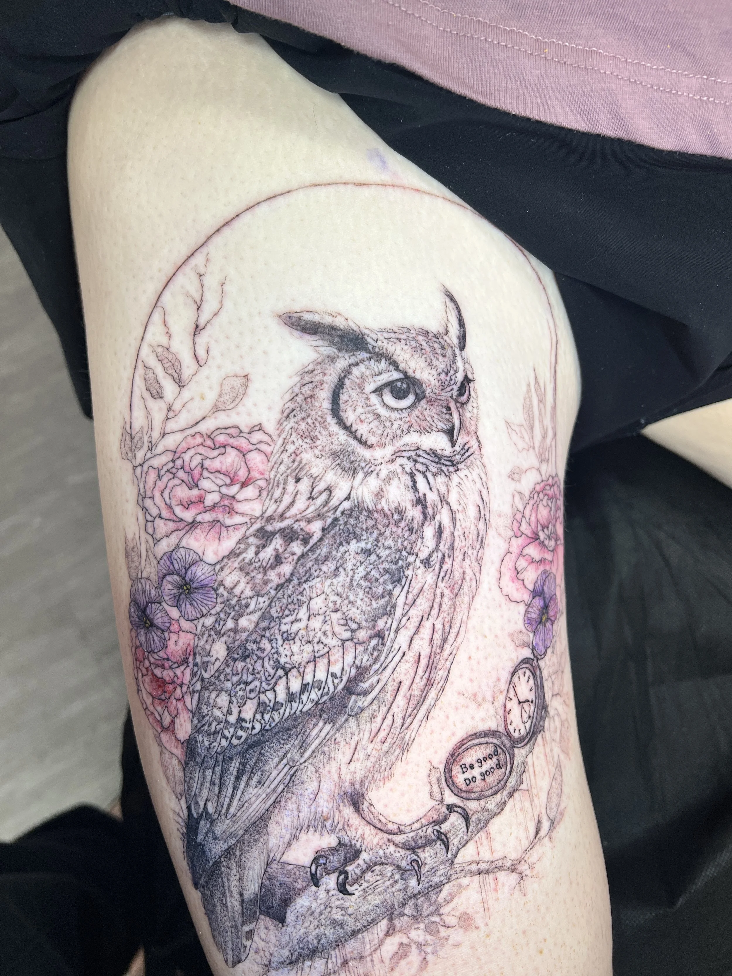 A tattoo of an owl surrounded by pink and purple flowers on a person's upper arm.