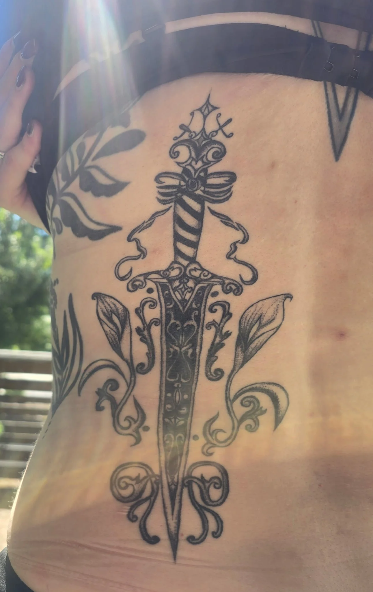 Tattoo of a decorative sword with ornate details on a person's back, surrounded by floral patterns.