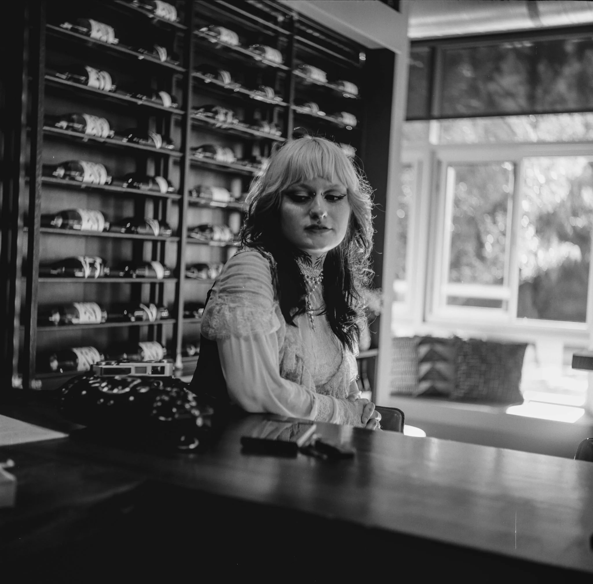 A woman with blonde and black curly hair, wearing a lace top, is sitting at a table in a room with large windows and a bookshelf filled with various items, including stacked plates.