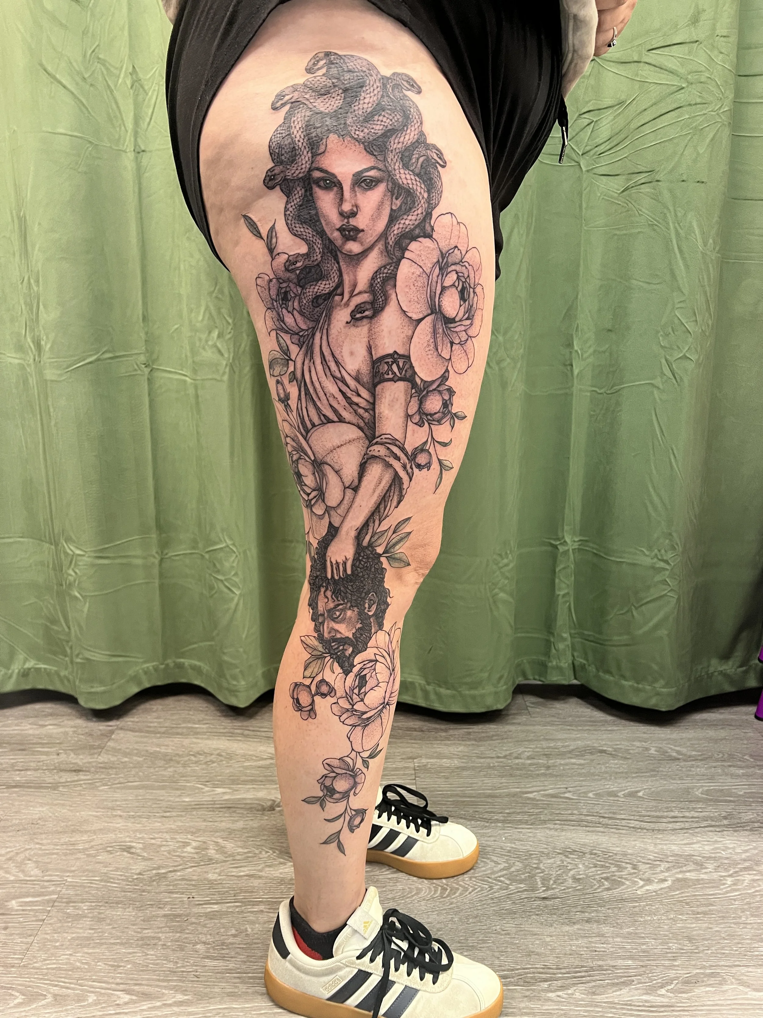 Tattoo of a woman with snakes as hair, surrounded by flowers, on a person's thigh, with the person wearing black shorts, sneakers, and standing on a wooden floor with a green backdrop.