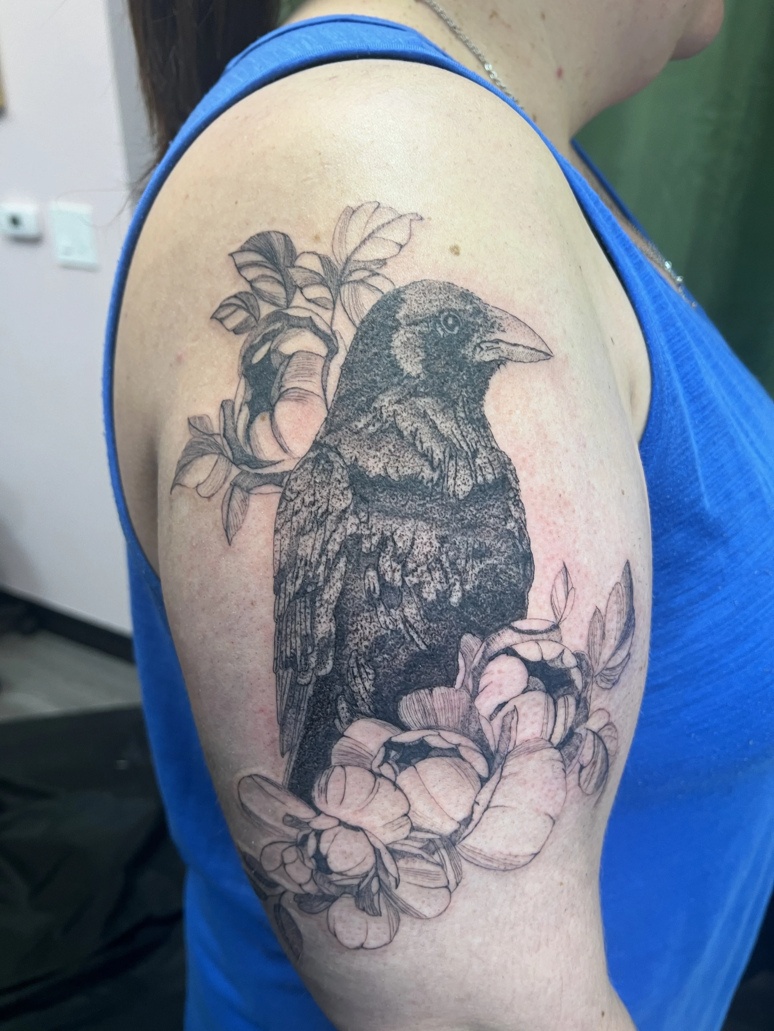A tattoo of a bird, likely a crow or raven, perched on a branch with leaves and flowers, inked in black and gray ink on a person's upper arm.