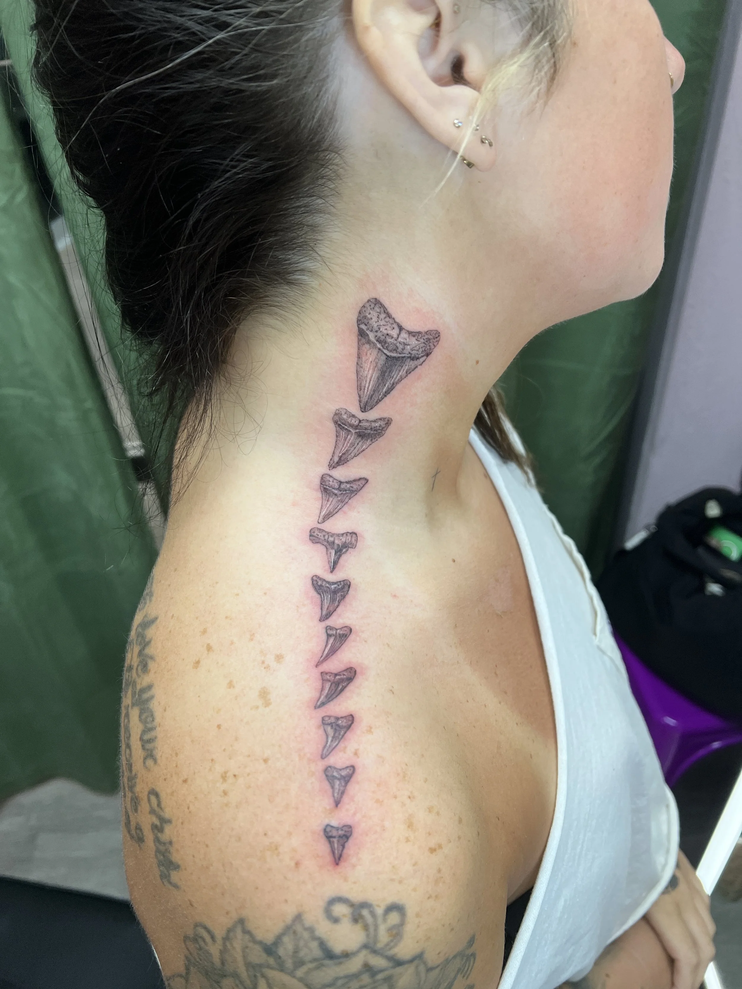 A woman with multiple ear piercings and dark hair pulled back, sitting in a chair. She has tattoos on her neck and upper arm; a tattoo on her neck features five fish skeleton designs vertically aligned.