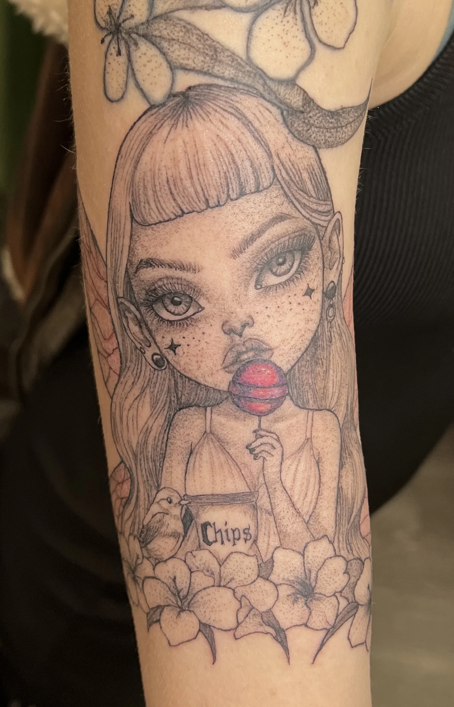 Tattoo of a stylized woman with big eyes, freckles, and earrings, holding a box of chips. The tattoo features realistic details with flowers and leaves surrounding the woman.