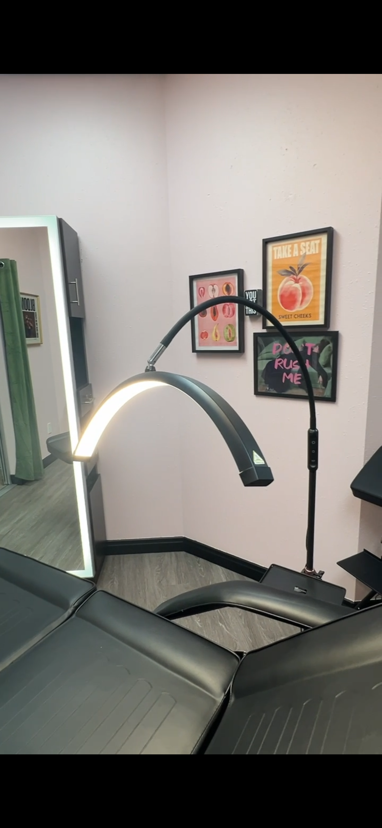 A tattoo chair with a curved black arm and a bright light, located in a room with pink walls. On the wall, three framed art pieces are visible: one with various fruits, another with a peach and the text 'Take a Seat', and the third with the phrase 'Don’t Ruin Me' over a leafy background.