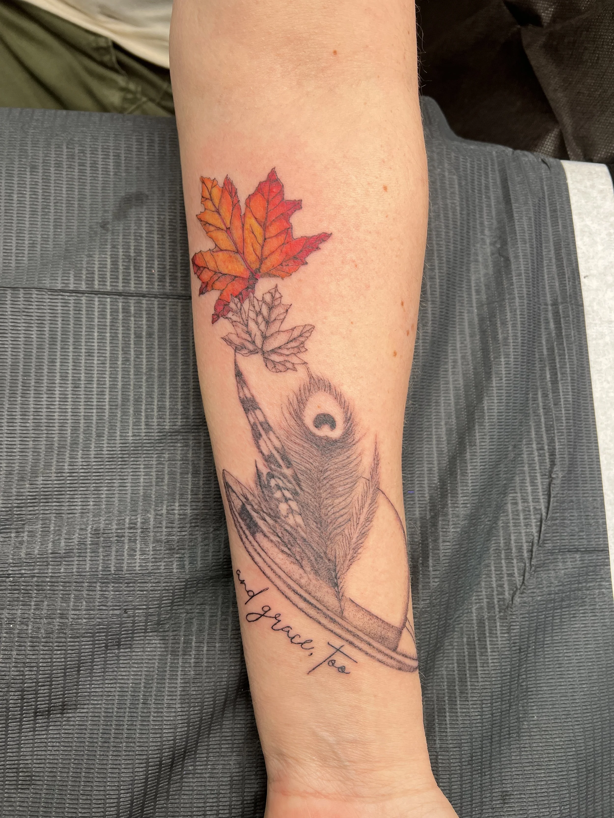 A person's forearm with a tattoo of a colorful autumn leaf and the phrase "Grace, To" written in cursive.