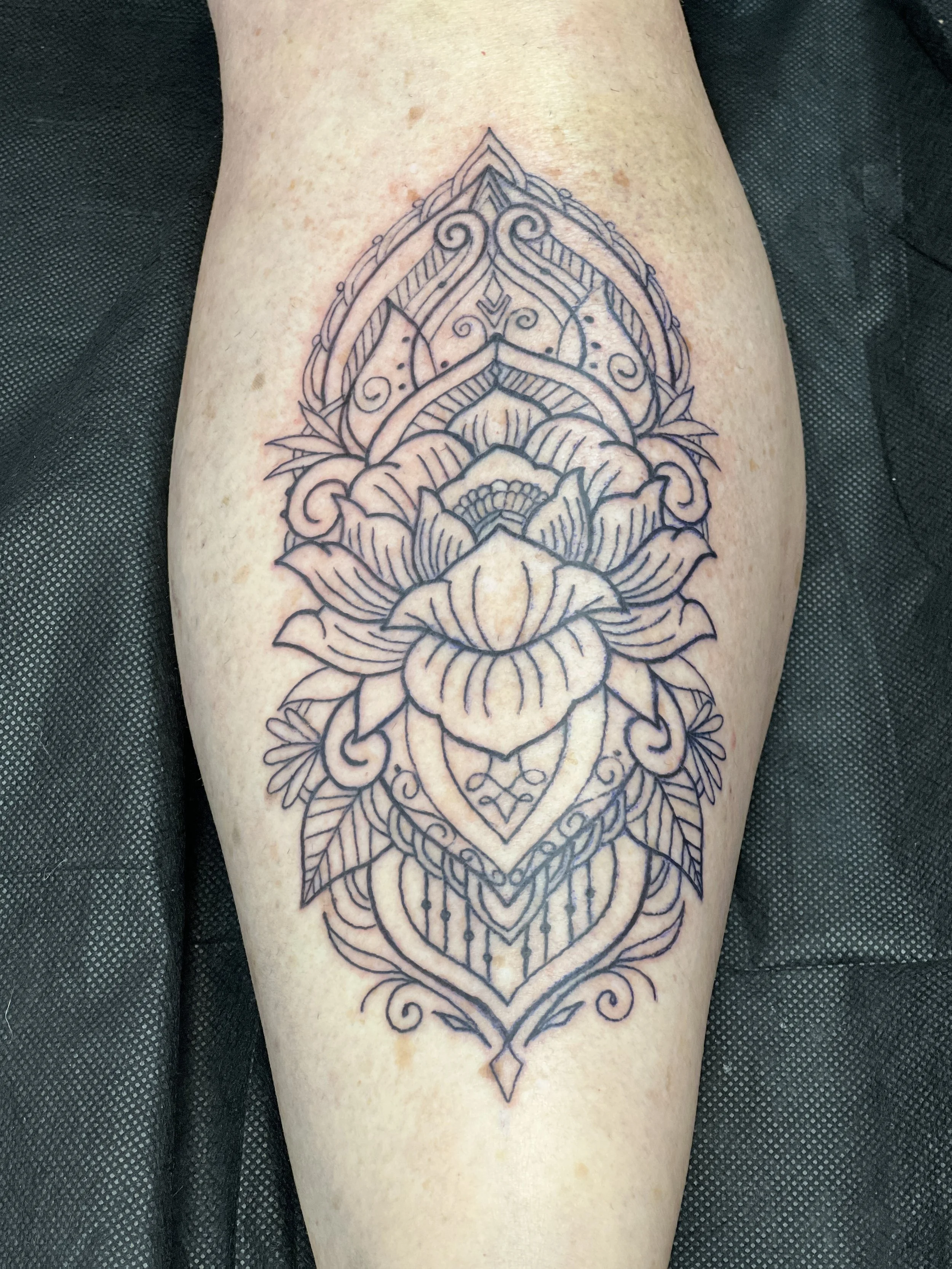 A detailed black ink line tattoo on a person's thigh featuring a symmetrical floral and geometric design, with intricate patterns and pointed shapes.