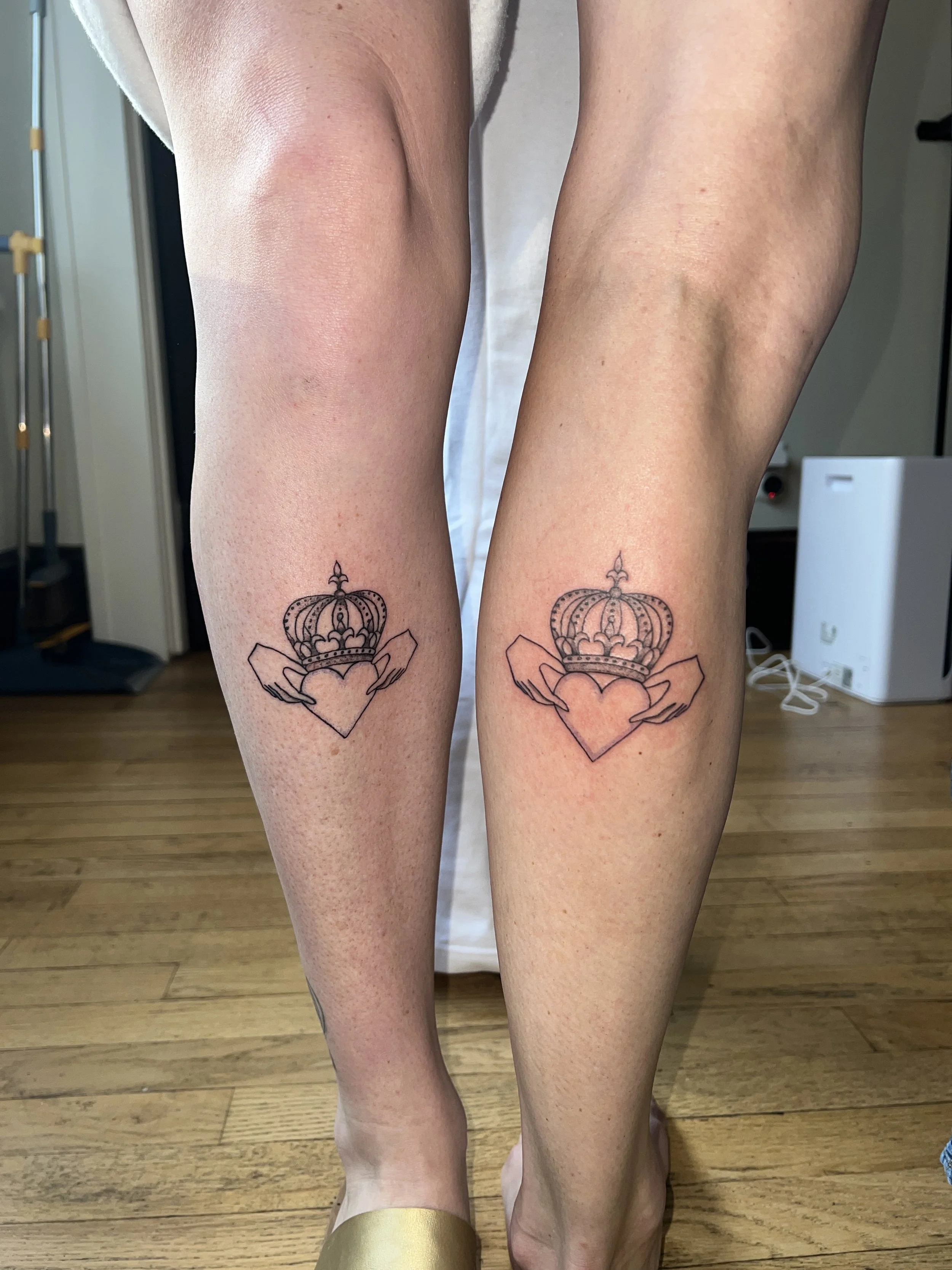 Two legs with matching tattoos of a crown and a heart, standing on a wooden floor.