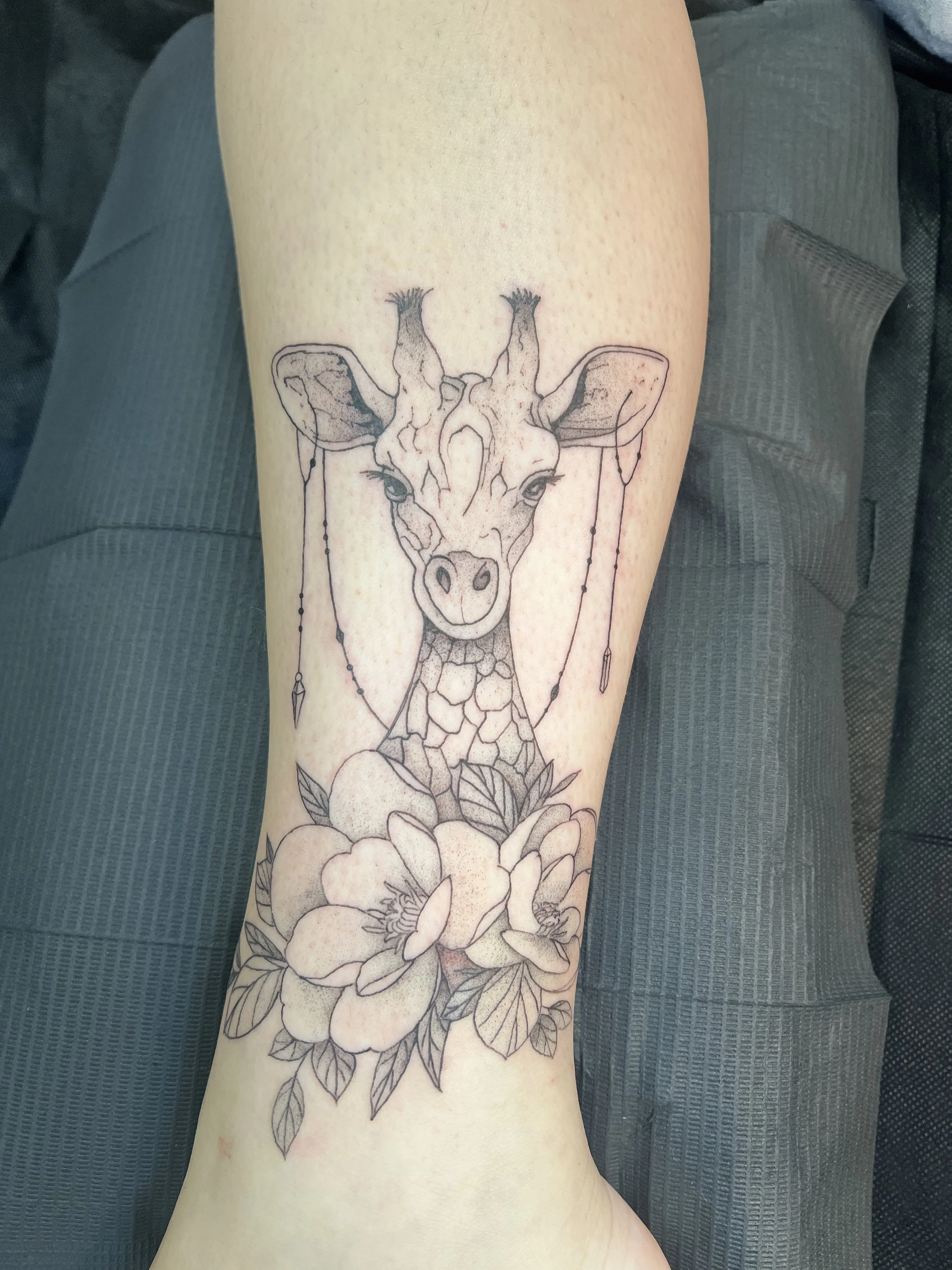 Black fine line art tattoo of a giraffe with a floral design below it on a person's forearm.