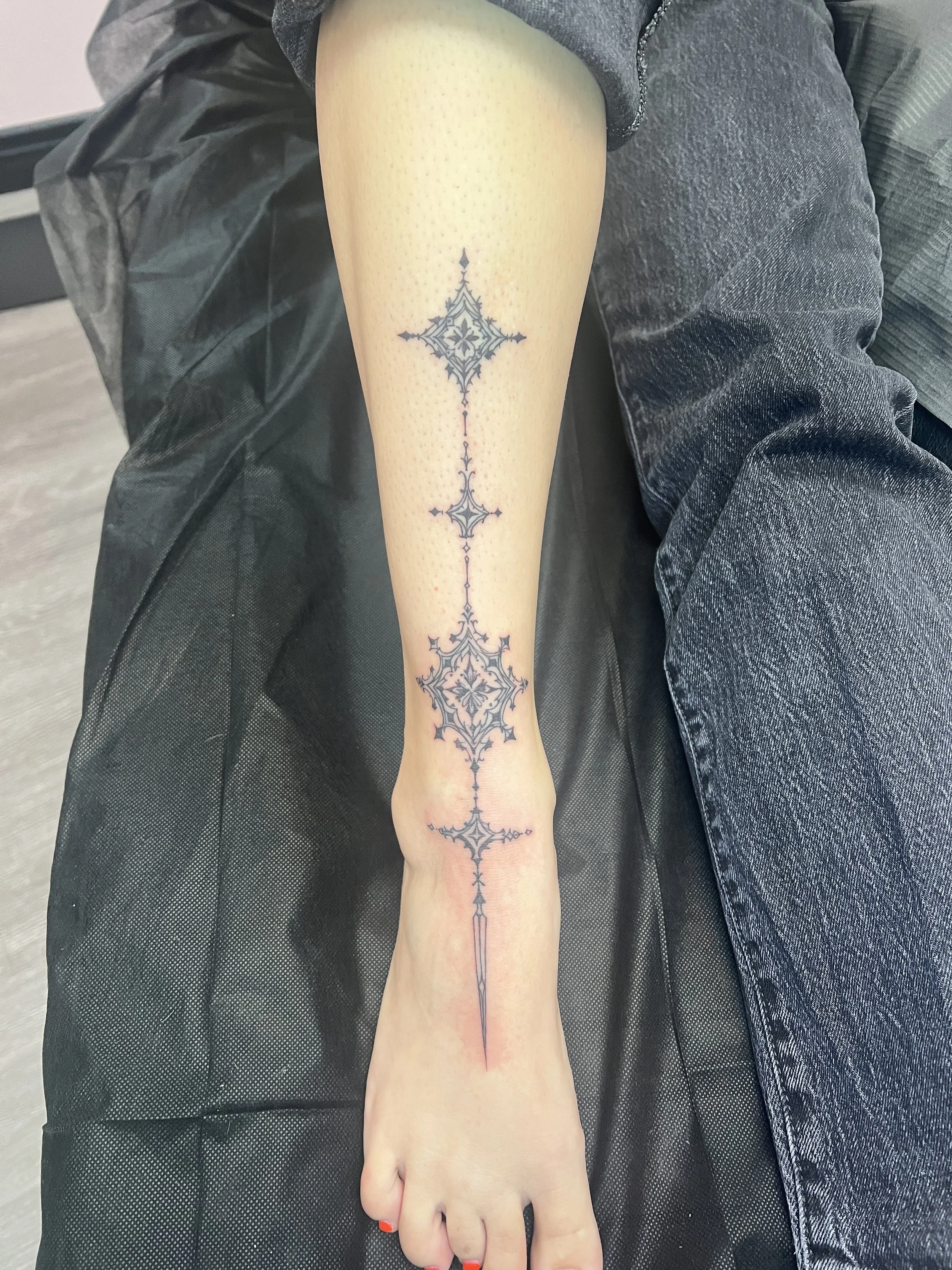 A person's leg with a detailed black ink tattoo running vertically along the front. The tattoo features symmetrical, intricate patterns and shapes that extend from just below the knee to the ankle. The person has red nail polish on their toes and is 