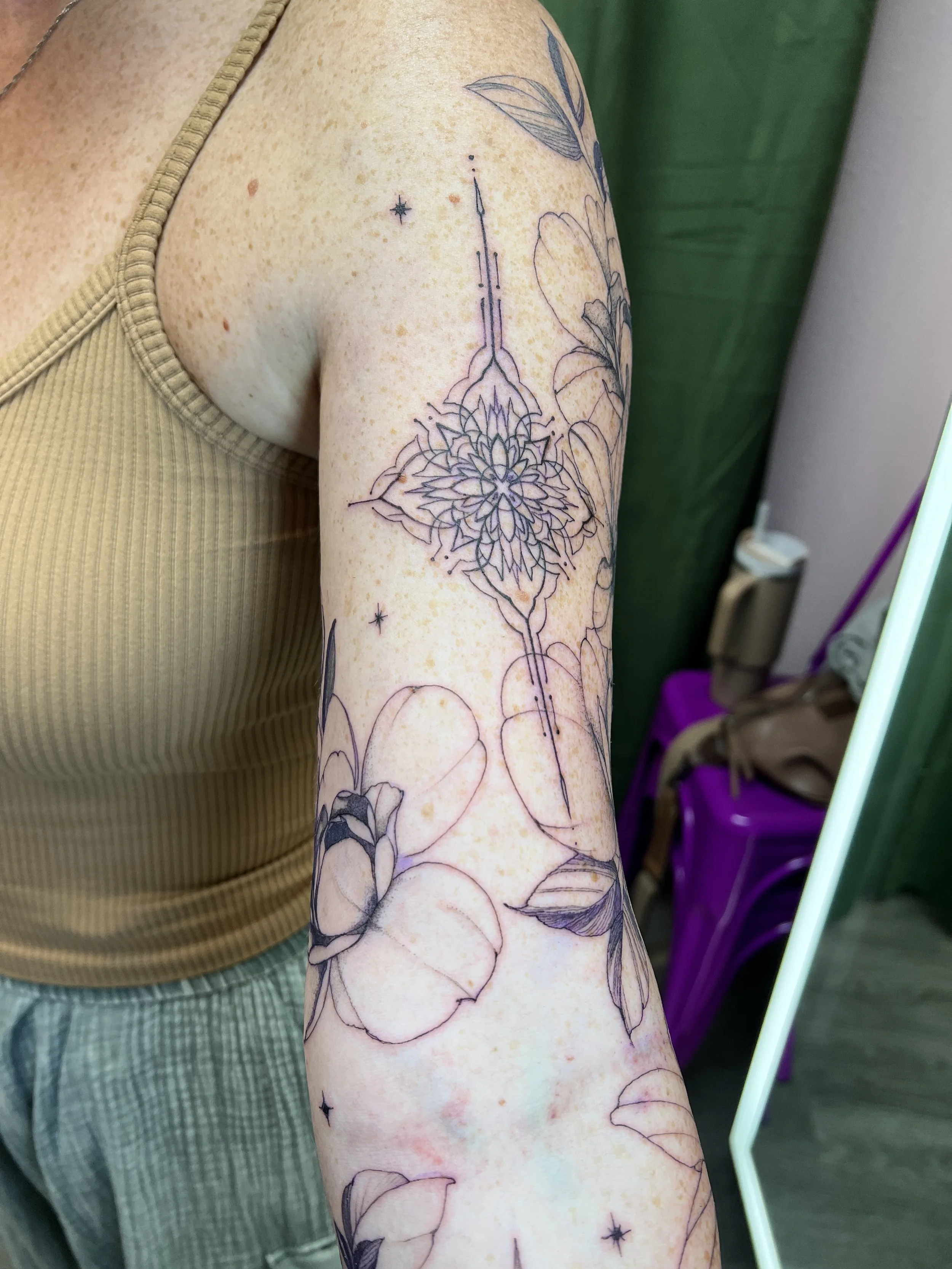 A person's arm with a floral tattoo in progress outlined in black ink, featuring flowers and leaves. The person is wearing a beige tank top and green checkered pants.