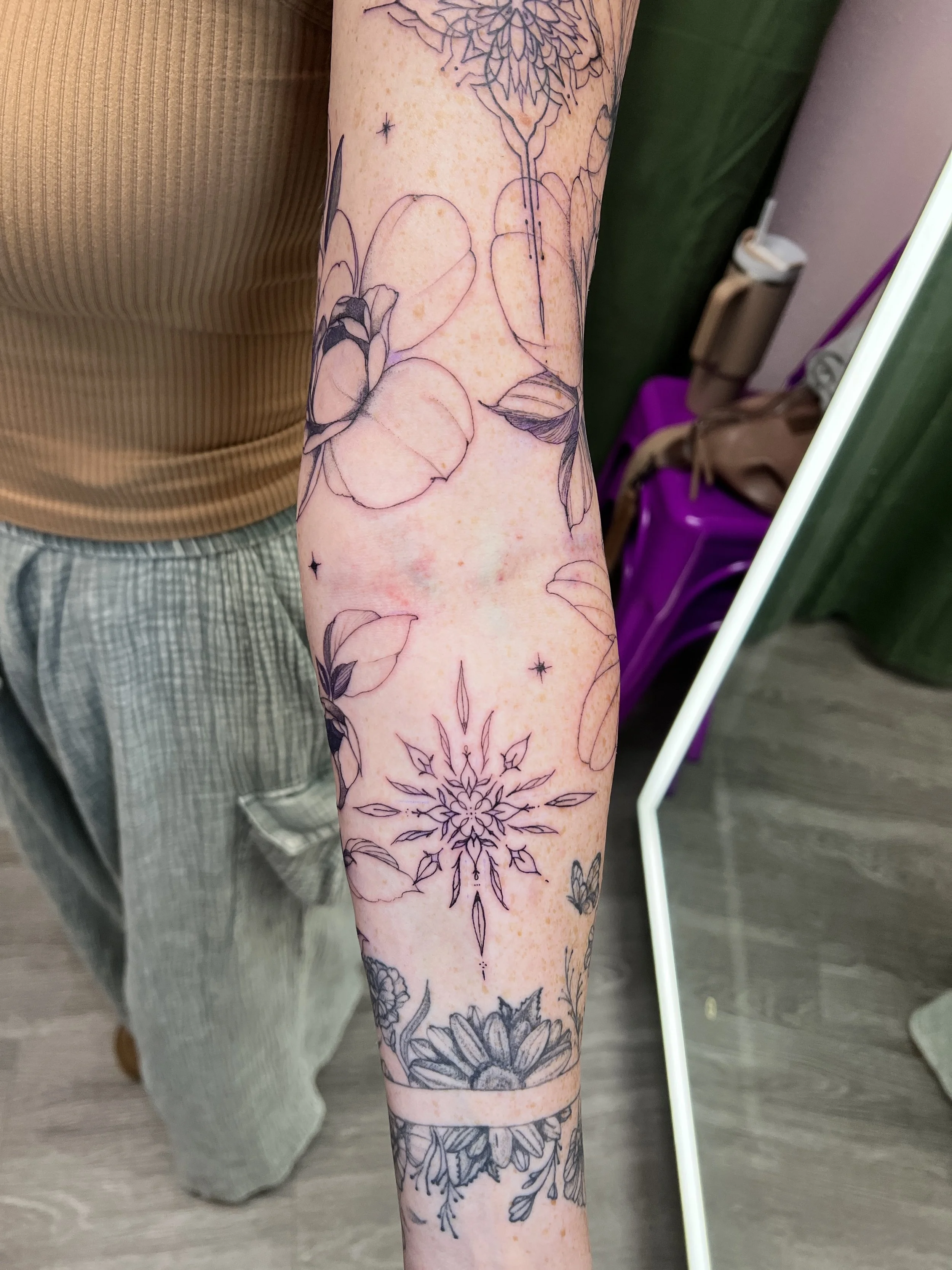 Close-up of a person's arm with multiple black outline floral tattoos, including different flowers and leaves, stretching from the wrist to the upper forearm. The person is wearing a light beige ribbed shirt and checkered shorts.