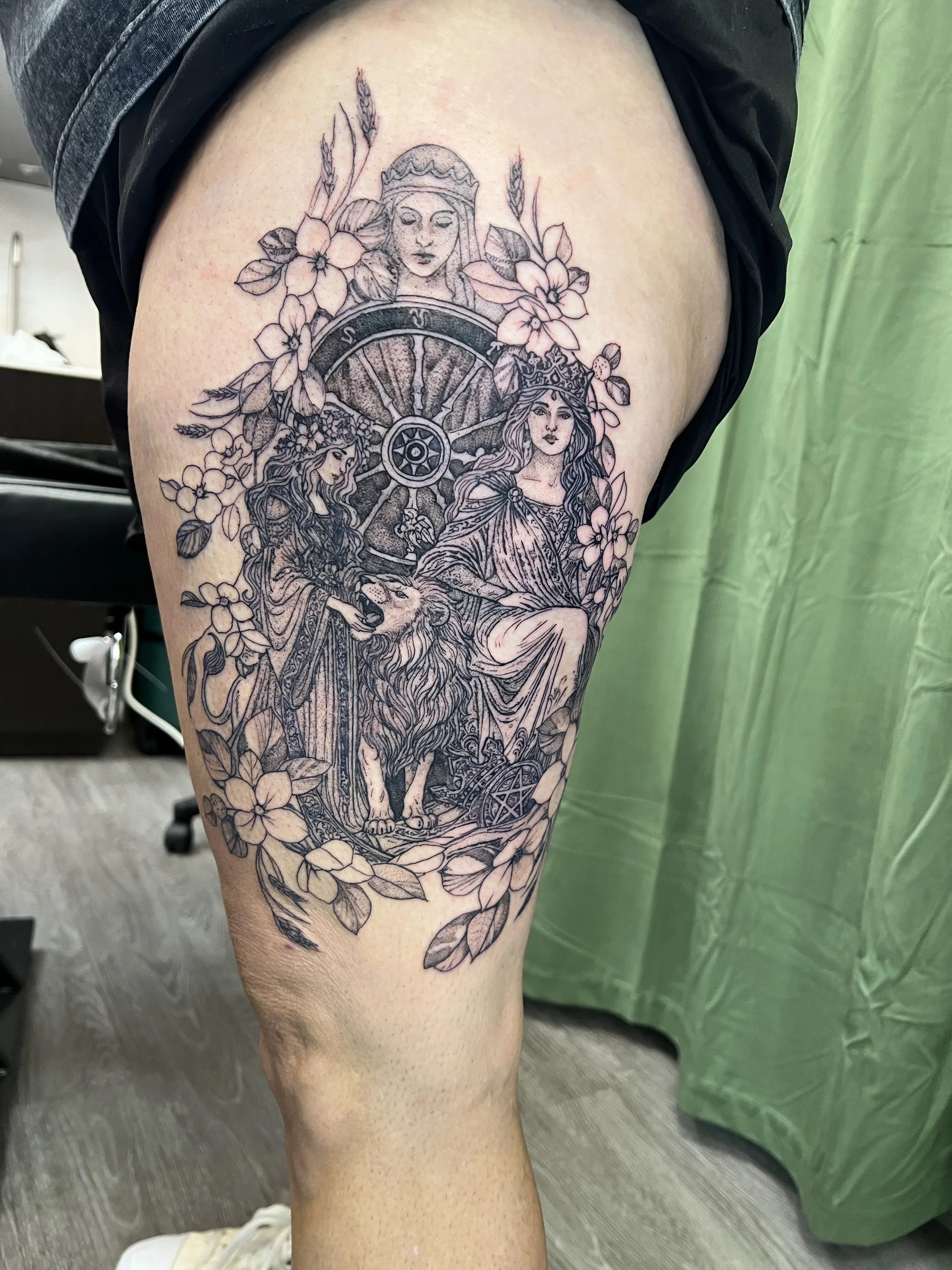 A detailed black and gray tattoo on a person's thigh featuring a woman's face wearing a crown, surrounded by flowers, leaves, and a wolf with a human-like face.
